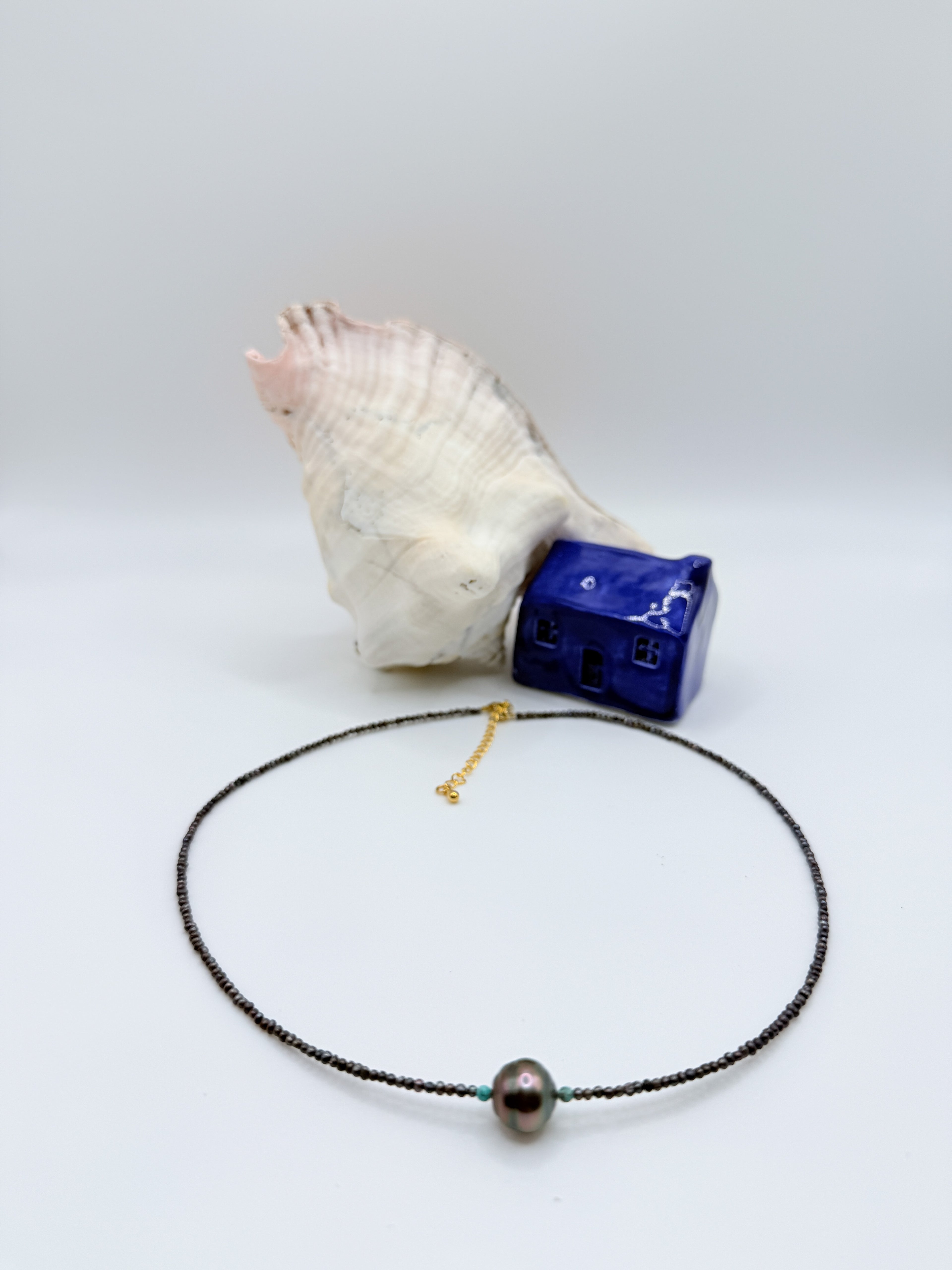 Minimalist choker with dark Tahitian baroque pearl and seed pearls displayed in circular formation with white conch shell and blue ceramic house on light background