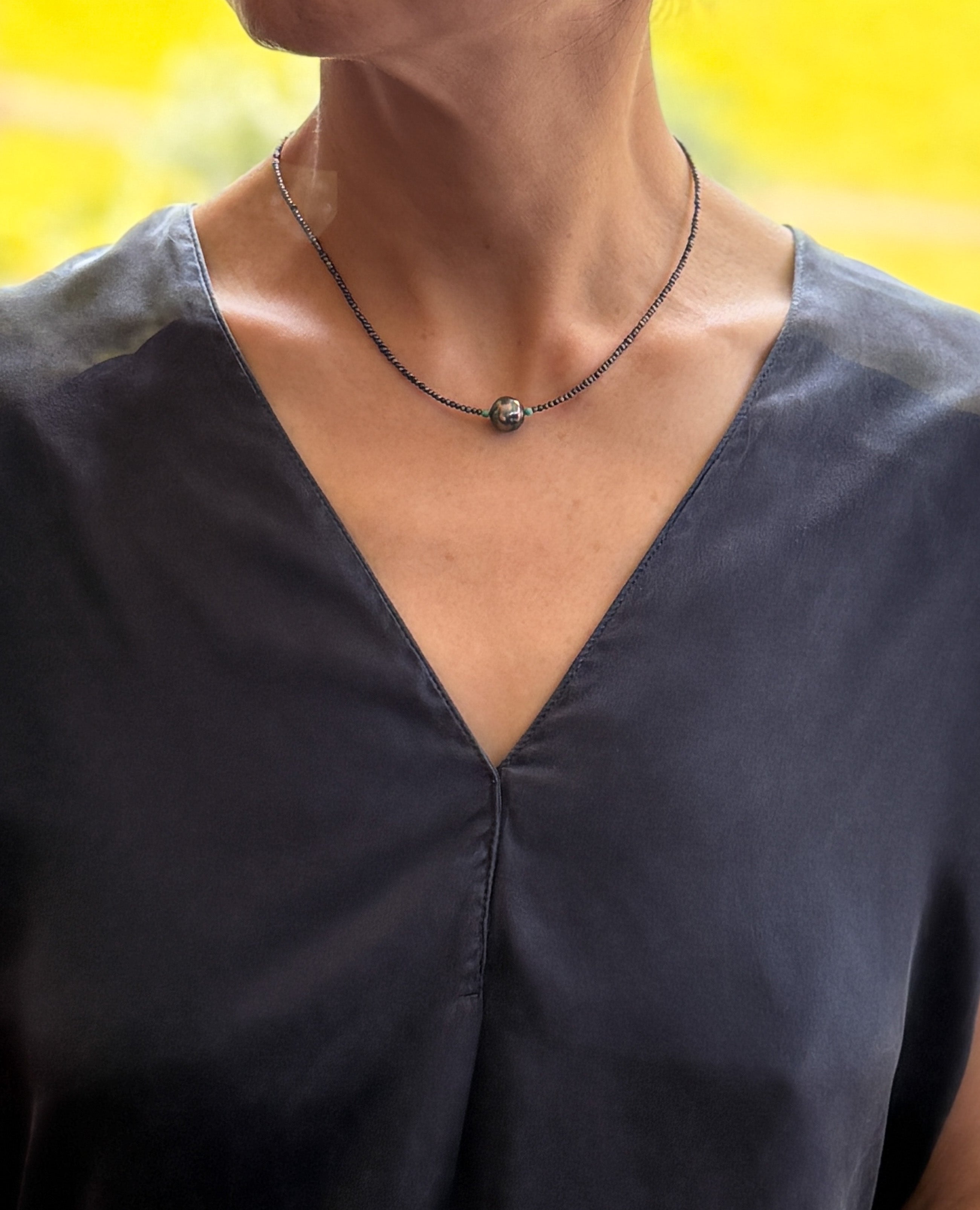 Model wearing delicate choker with dark Tahitian baroque pearl centrepiece and tiny seed pearl beads over navy-blue v-neck top with warm natural lighting
