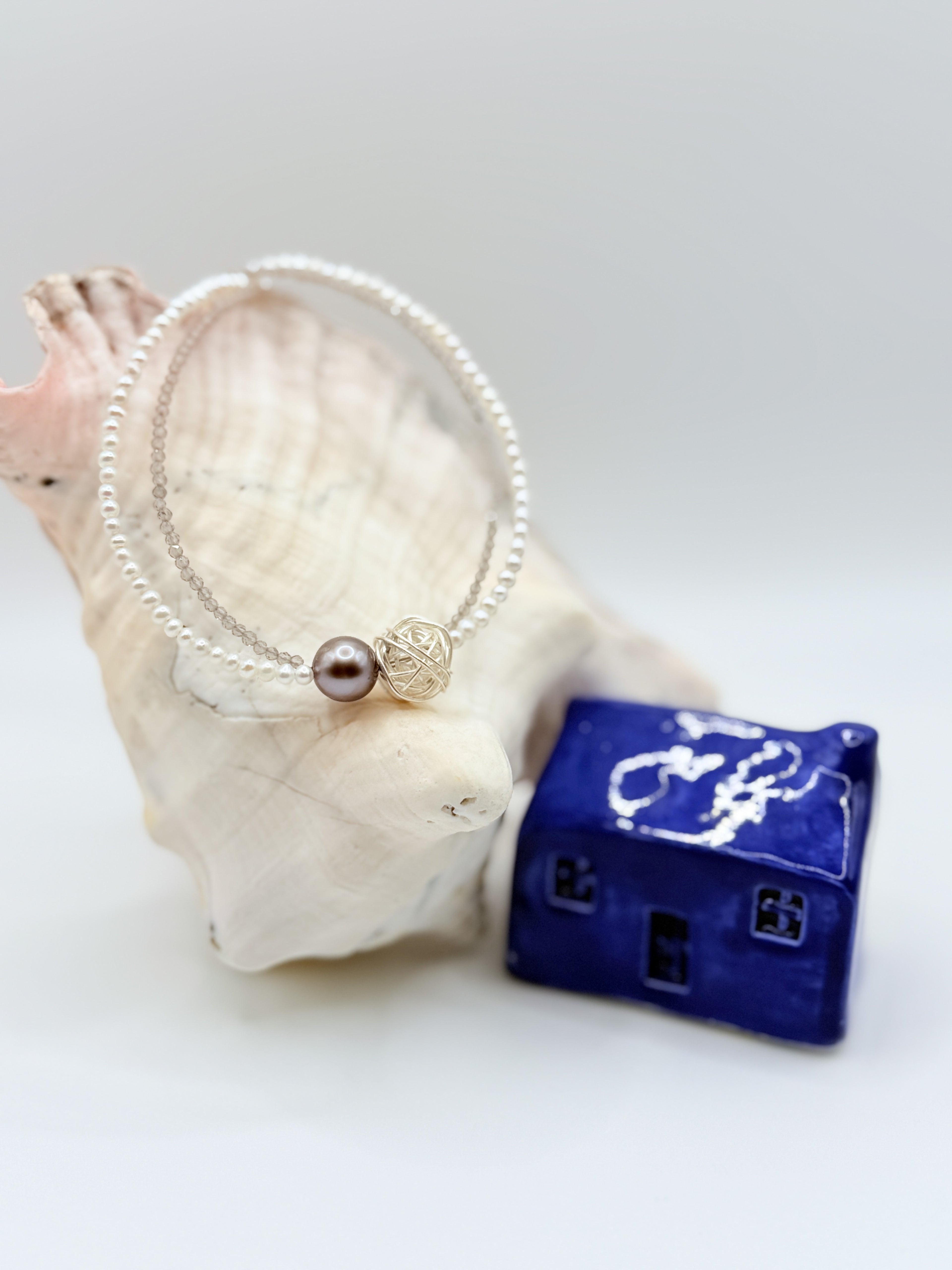 Delicate choker with dark Tahitian baroque pearl and silver wire cage bead surrounded by tiny white seed pearls, displayed in circular formation with white shell and blue ceramic house in soft focus