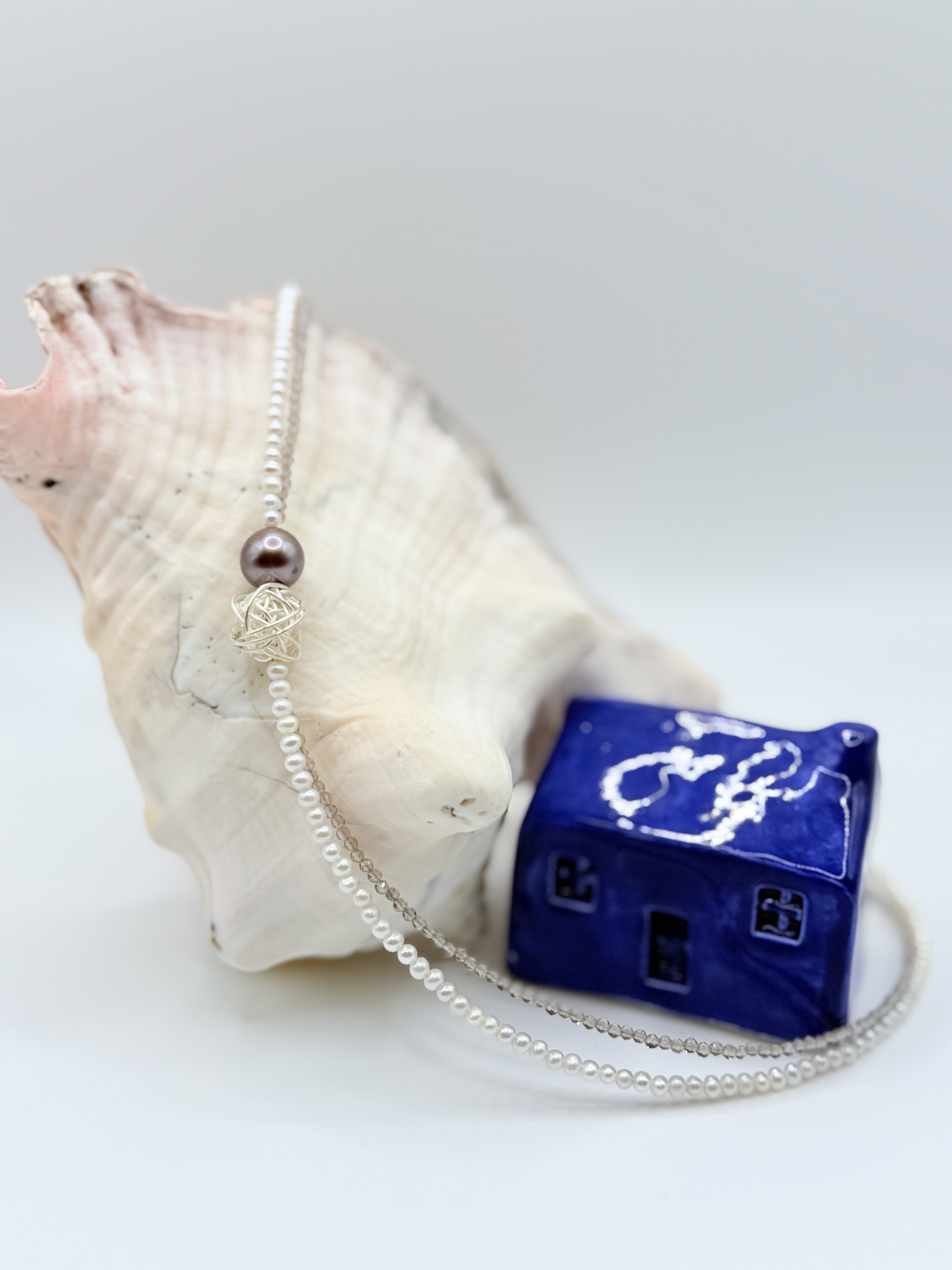Elegant choker with Tahitian baroque pearl and silver wire-wrapped bead flowing gracefully over white conch shell, showing the full strand of tiny white seed pearls with blue ceramic house