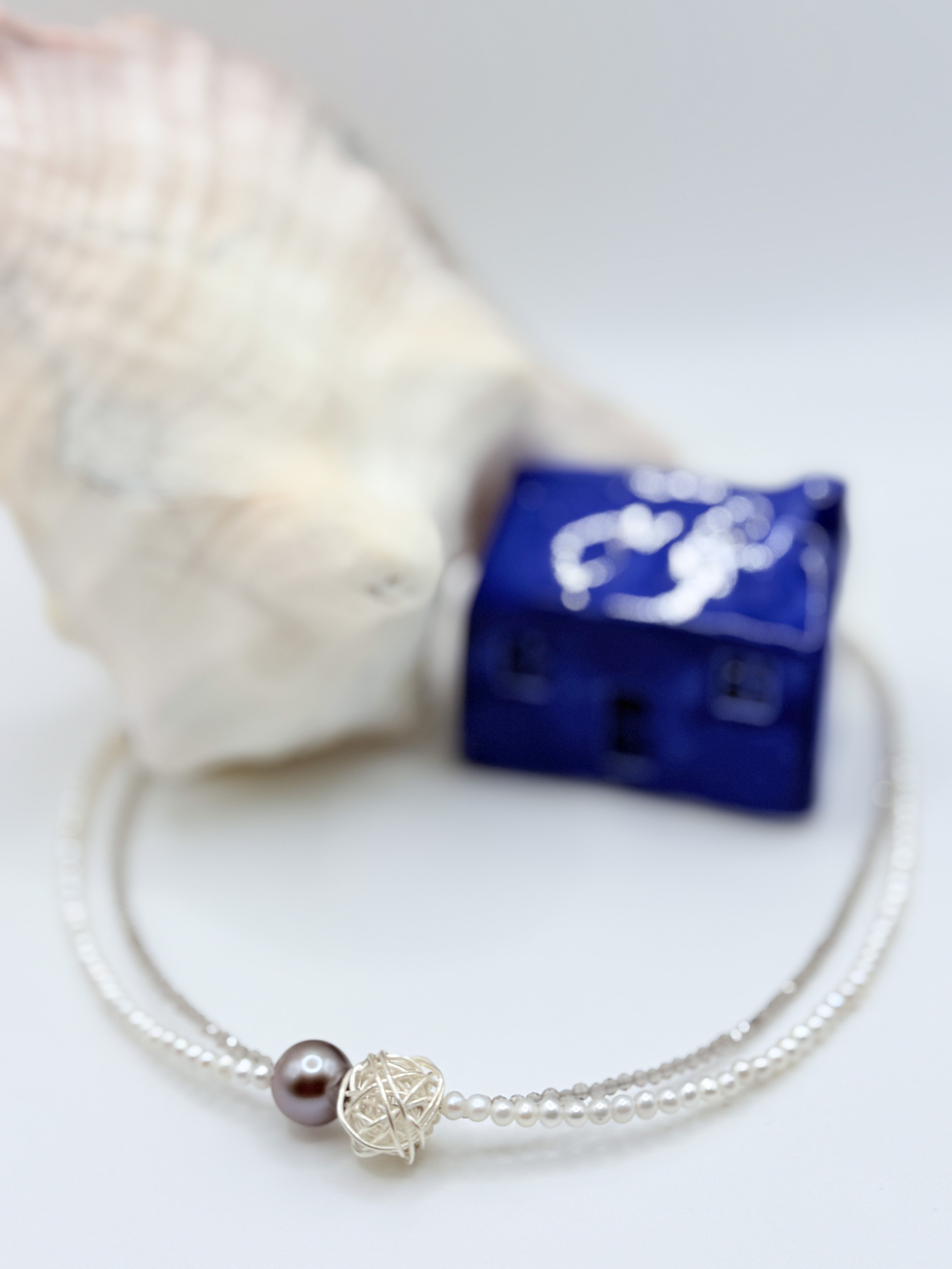 Close-up of elegant choker displayed in circular formation on white conch shell, highlighting the contrast between dark Tahitian baroque pearl, intricate silver wire-wrapped bead, and luminous white seed pearls