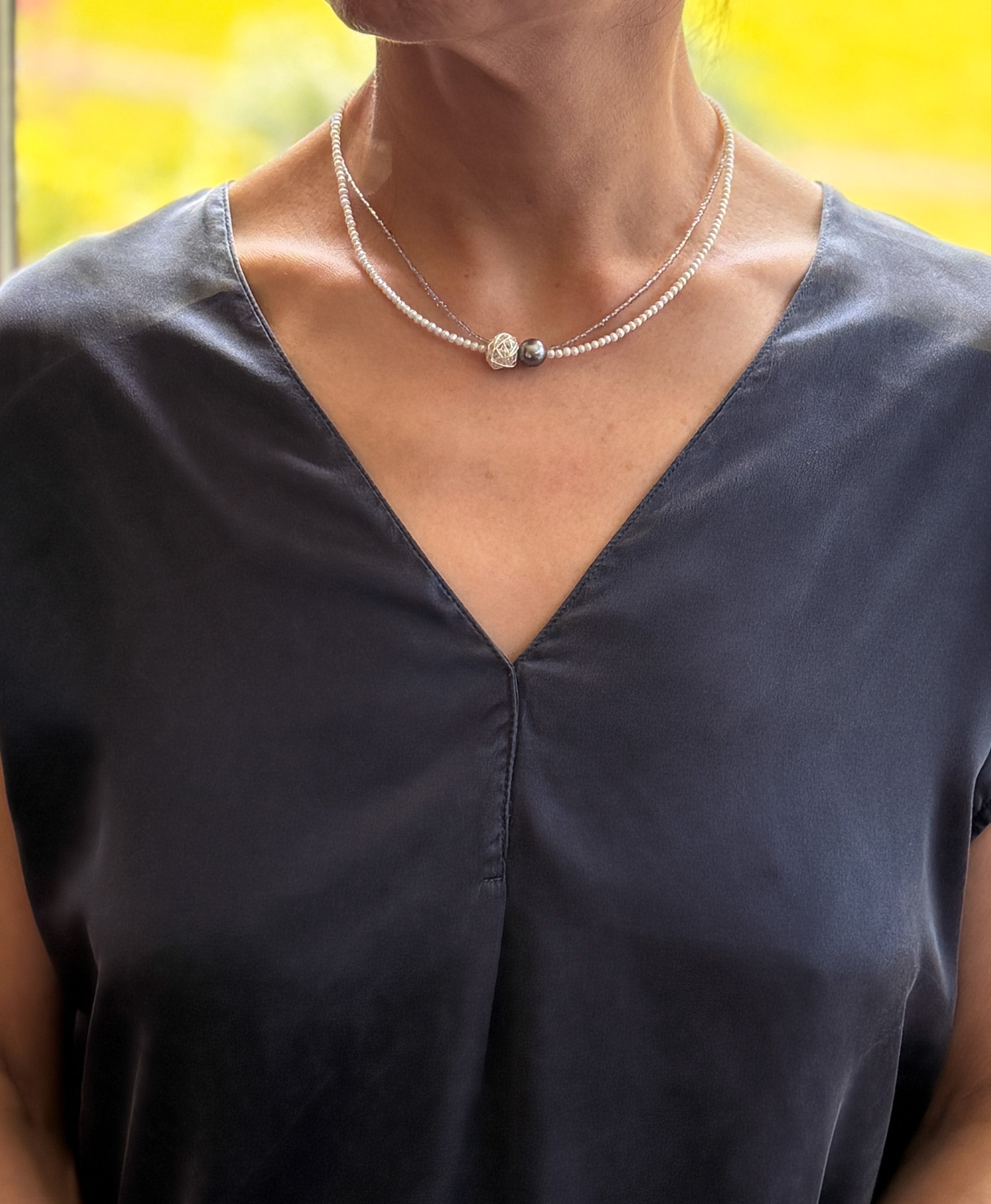 Model wearing elegant choker with Tahitian baroque pearl, silver wire-wrapped bead, and white seed pearls over navy-blue v-neck top with warm natural lighting