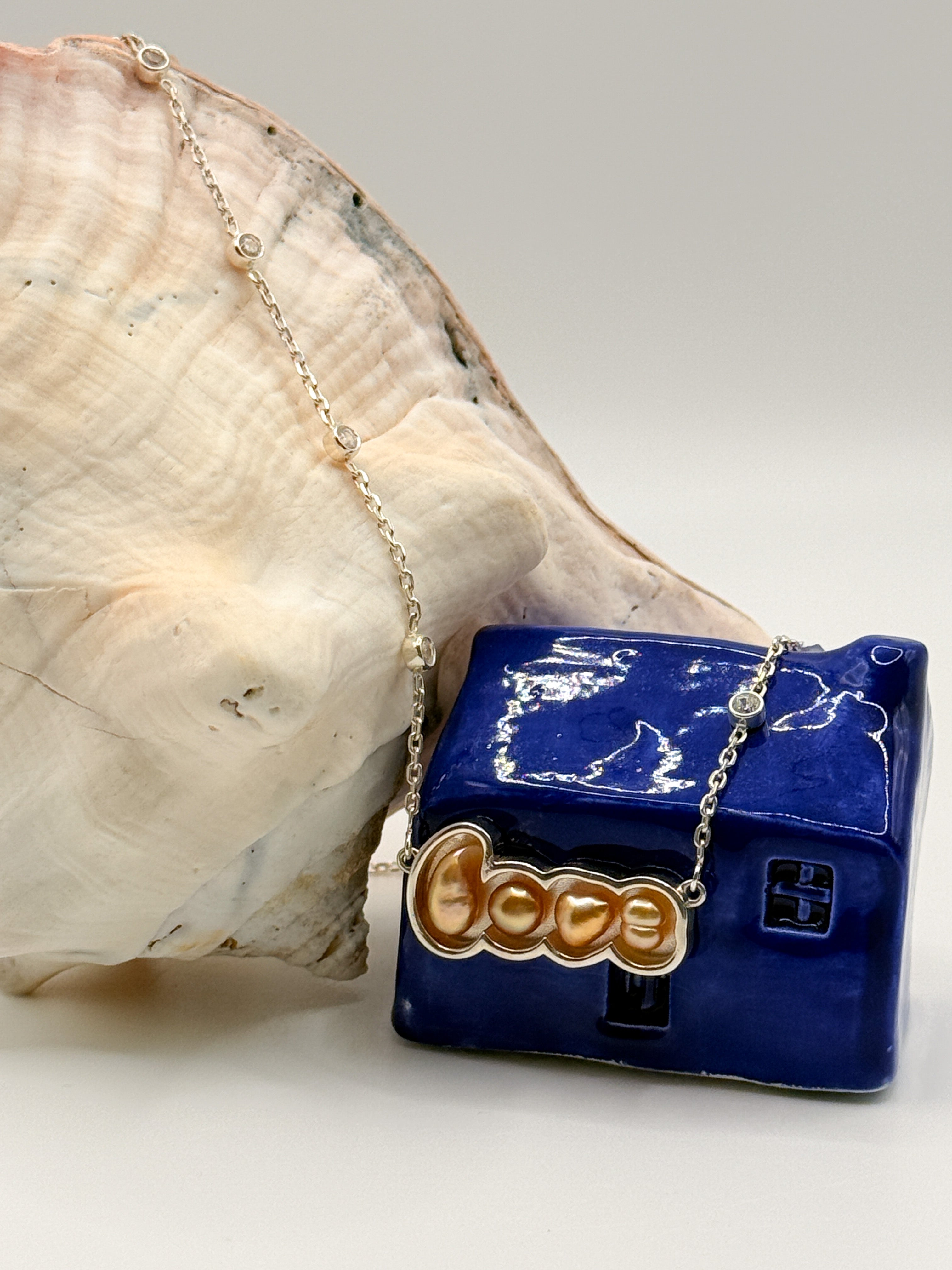 Gold 'LOVE' pendant necklace with South Sea keshi pearls draped over conch shell and blue ceramic house, showing the full chain length with CZ station details