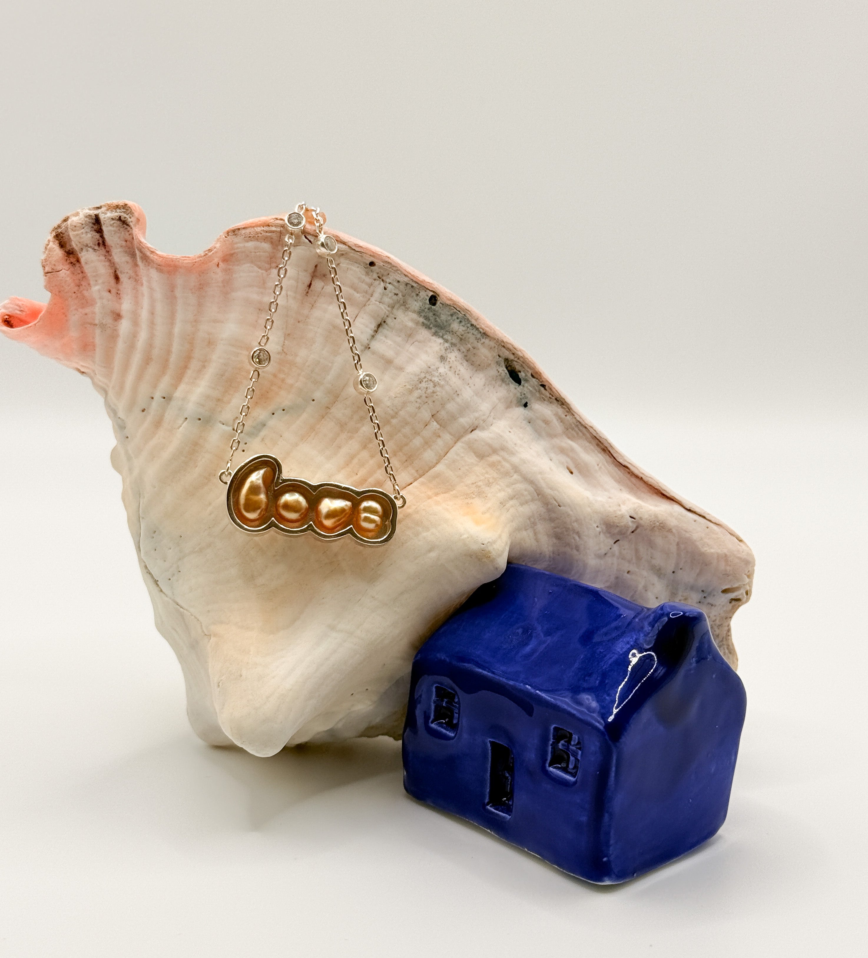 Gold 'LOVE' pendant necklace with South Sea keshi pearls displayed on large conch shell next to blue ceramic house, showing the word spelled out in connected pearl settings