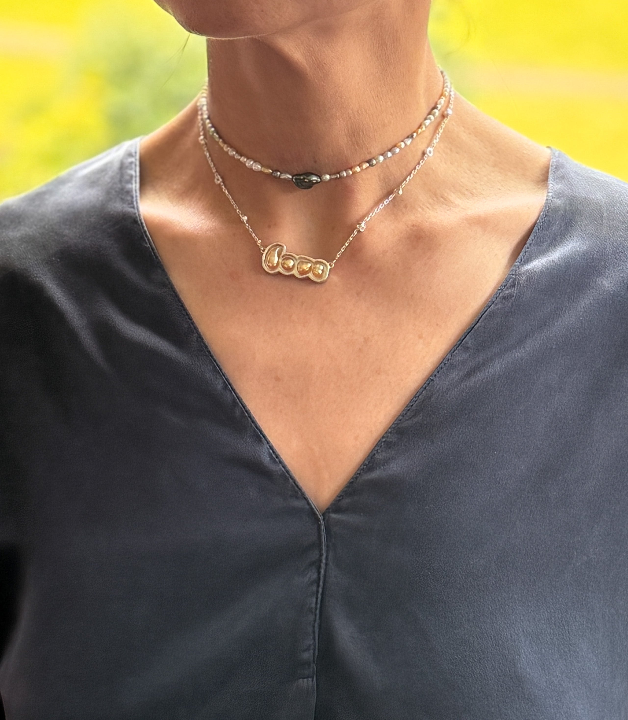Model wearing layered necklaces including gold 'LOVE' pendant with South Sea keshi pearls and seed pearl choker over charcoal grey v-neck top with warm natural lighting