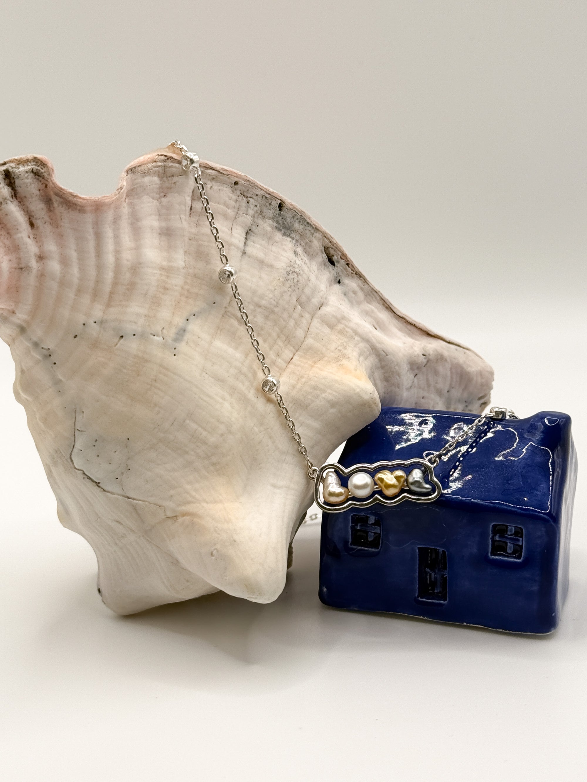 Sterling silver 'LOVE' pendant necklace with mixed South Sea and Tahitian keshi pearls draped over conch shell and blue ceramic house, highlighting the diverse pearl tones