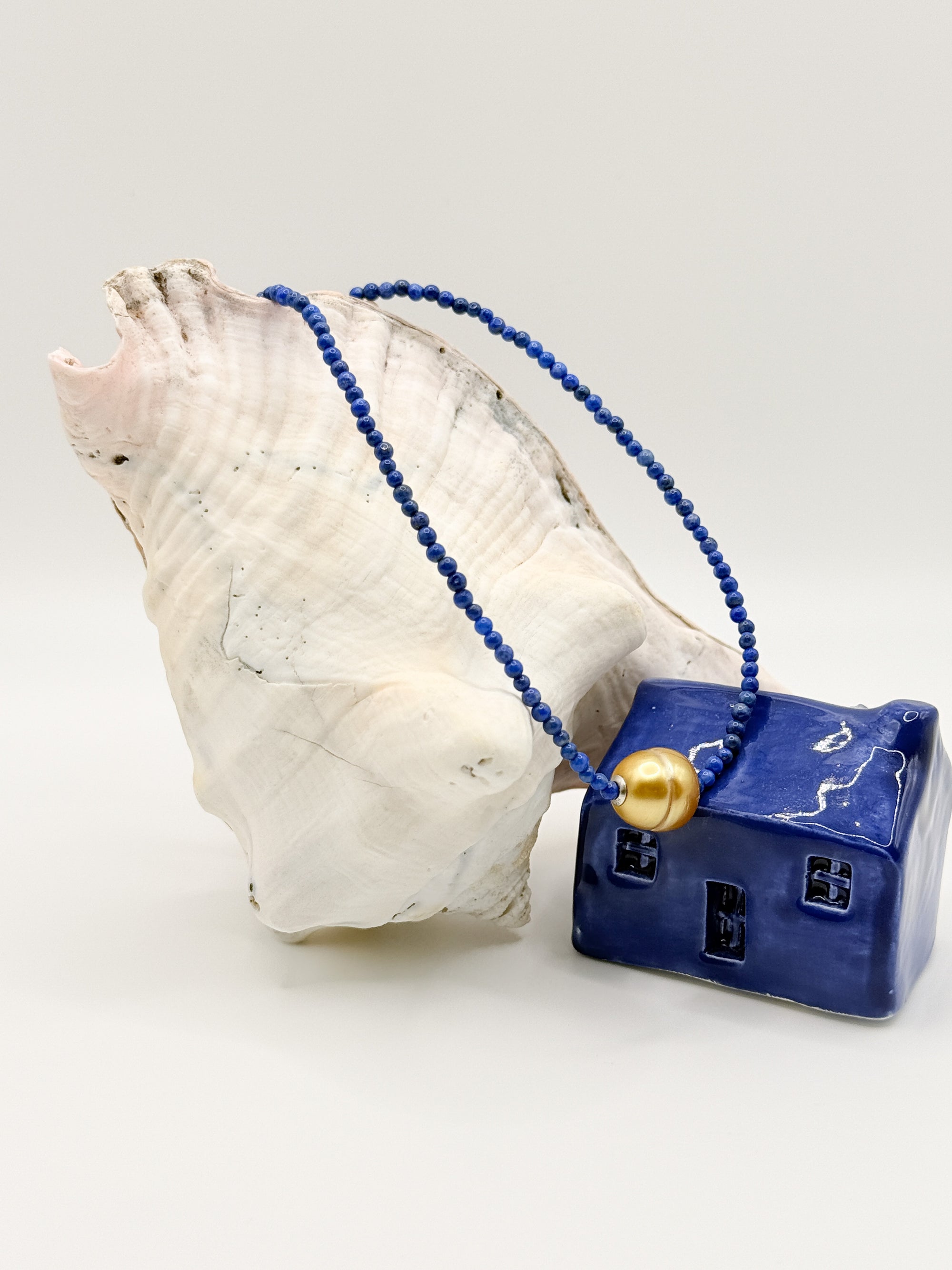 Gold South Sea pearl choker with deep blue lapis lazuli beads draped over white conch shell next to blue ceramic house, showing the golden pearl centrepiece