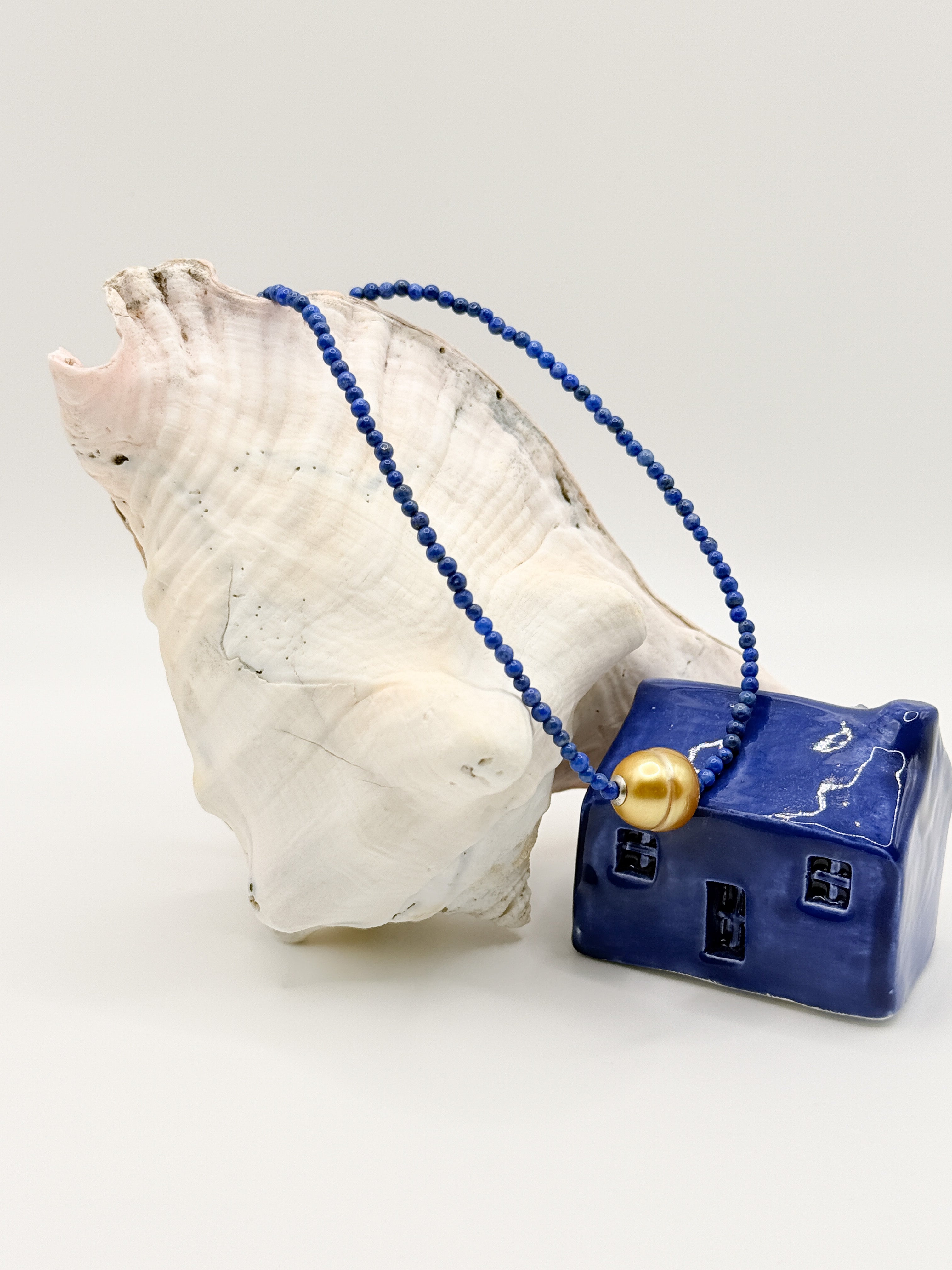 Gold South Sea pearl choker with deep blue lapis lazuli beads draped over white conch shell next to blue ceramic house, showing the golden pearl centrepiece