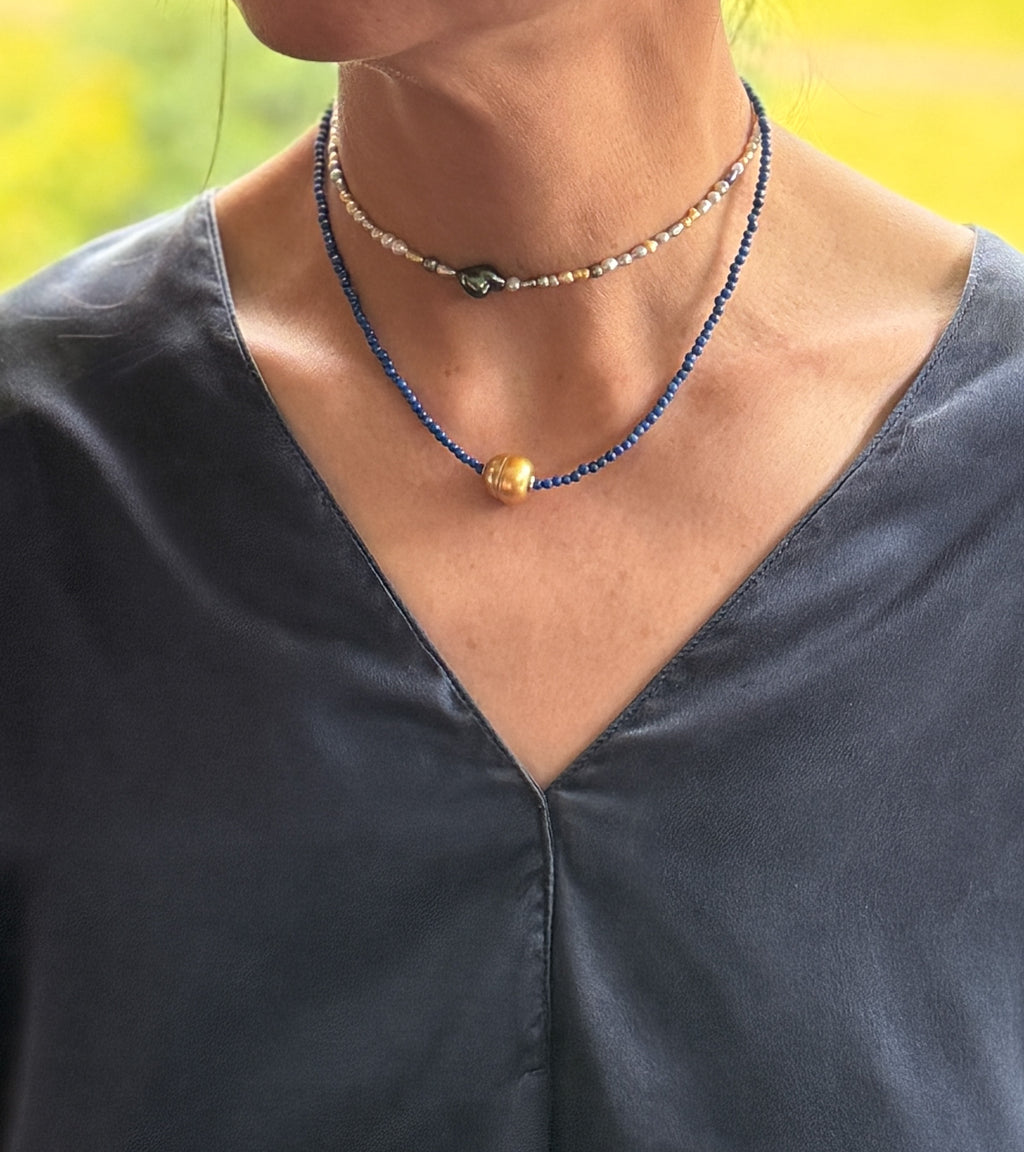 Model wearing layered necklaces including golden South Sea pearl choker with lapis lazuli beads and seed pearl choker over navy-blue v-neck top with warm natural lighting