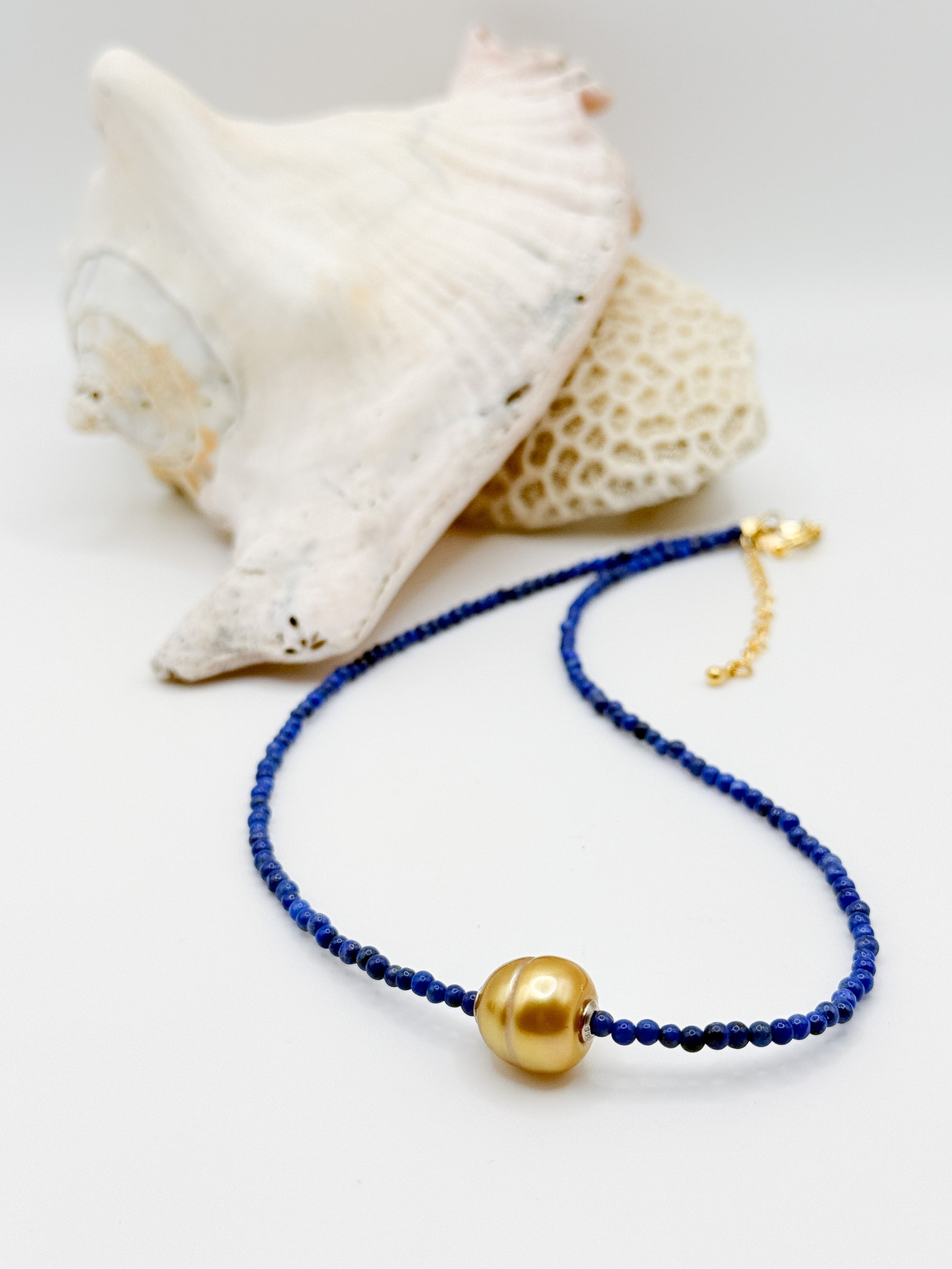 Golden South Sea pearl choker with lapis lazuli beads laid in curved formation next to white coral specimen, showcasing the contrast between deep blue beads and lustrous golden pearl