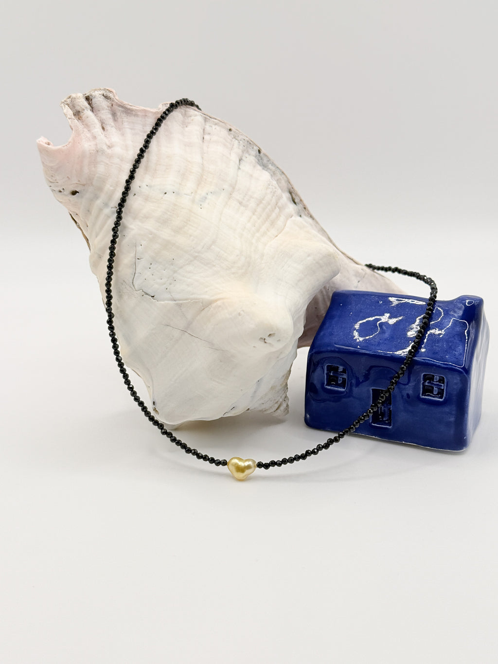 Golden South Sea heart keshi pearl choker with black spinel beads displayed in circular formation around white conch shell and blue ceramic house