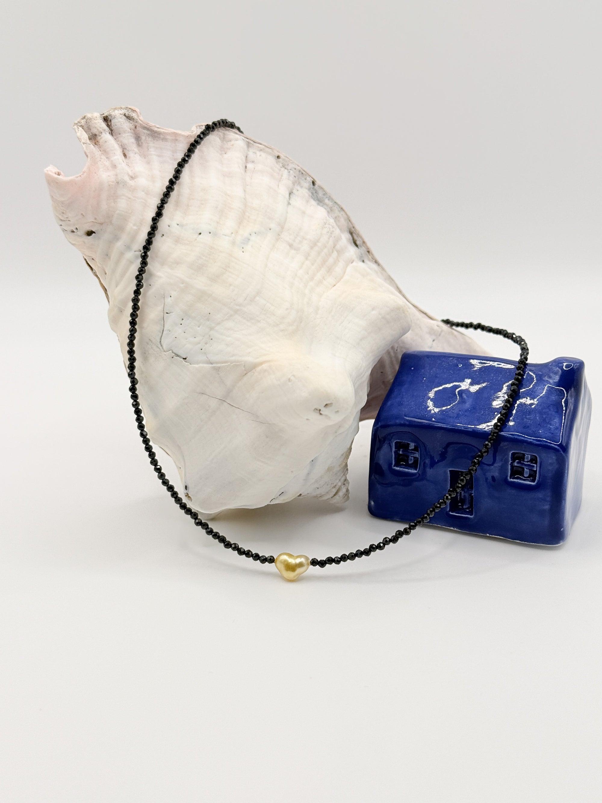 Golden South Sea heart keshi pearl choker with black spinel beads displayed in circular formation around white conch shell and blue ceramic house
