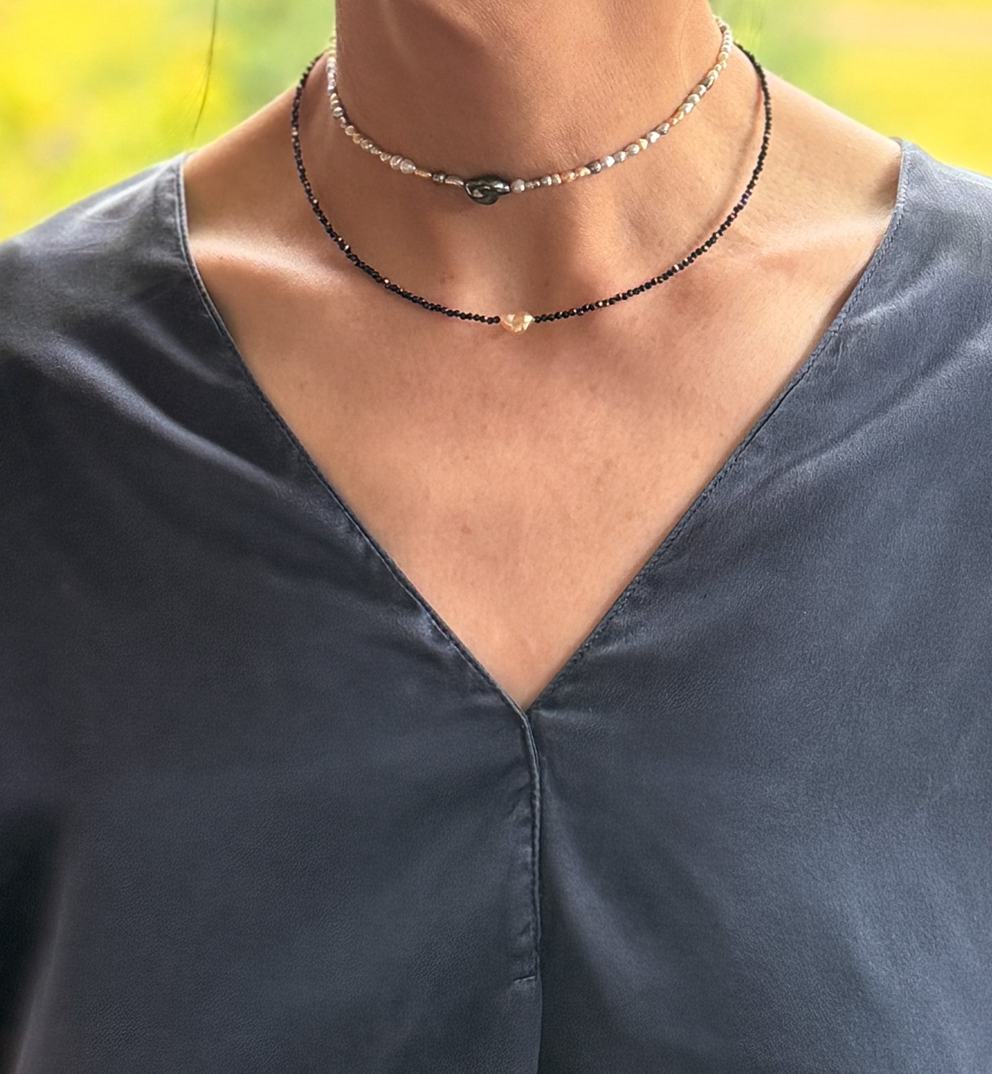 Model wearing layered necklaces including golden South Sea heart keshi pearl choker with black spinel beads and seed pearl choker over navy blue v-neck top with warm natural lighting