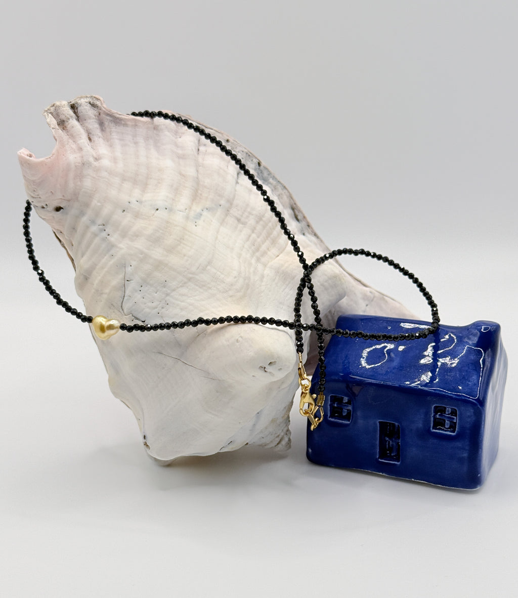 Golden South Sea heart keshi pearl choker with black spinel beads flowing gracefully over white conch shell and blue ceramic house, showing the full strand length with gold-coloured clasp details