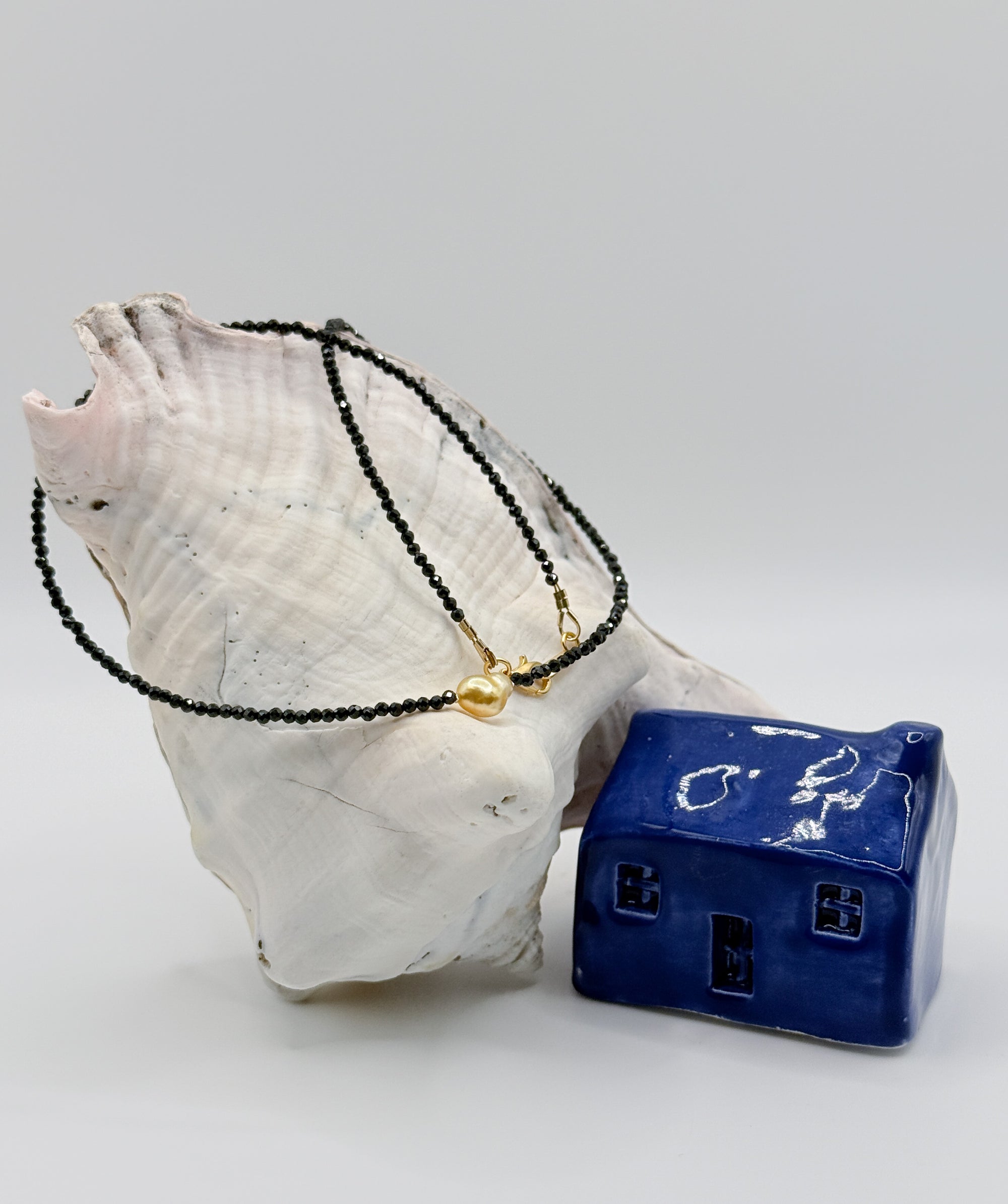Black spinel bead choker with golden South Sea heart-shaped keshi pearl centrepiece draped over white conch shell next to blue ceramic house
