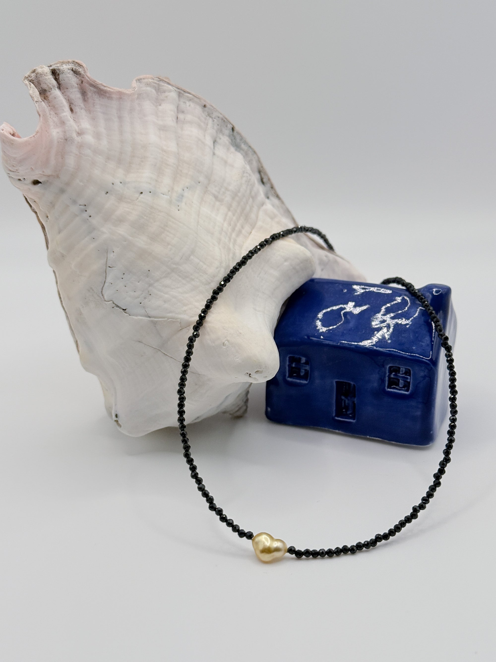 Golden South Sea heart keshi pearl choker with black spinel beads elegantly draped over white conch shell and blue ceramic house, highlighting the romantic heart-shaped pearl centrepiece