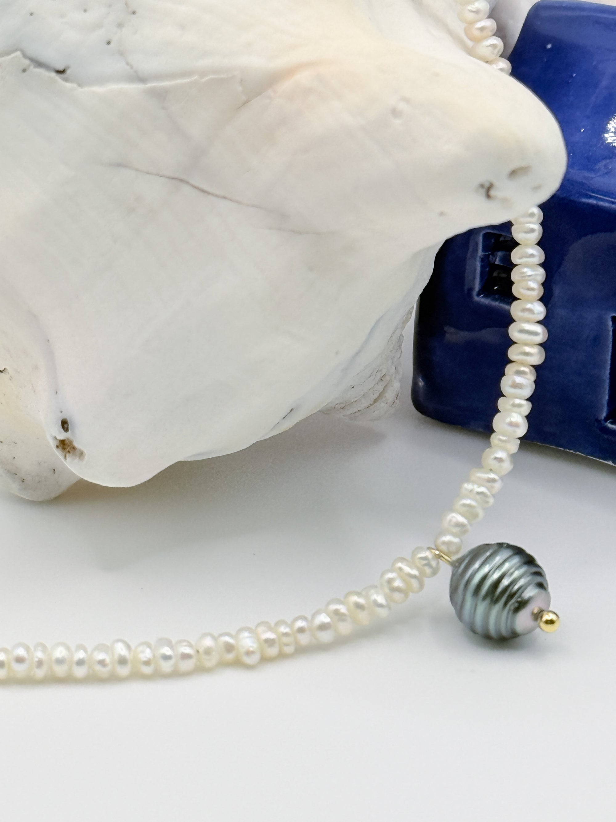 Close-up detail of white seed pearl choker with green Tahitian baroque circled pearl pendant flowing over white conch shell and blue ceramic house, showcasing the lustrous surface and texture of the pendant pearl