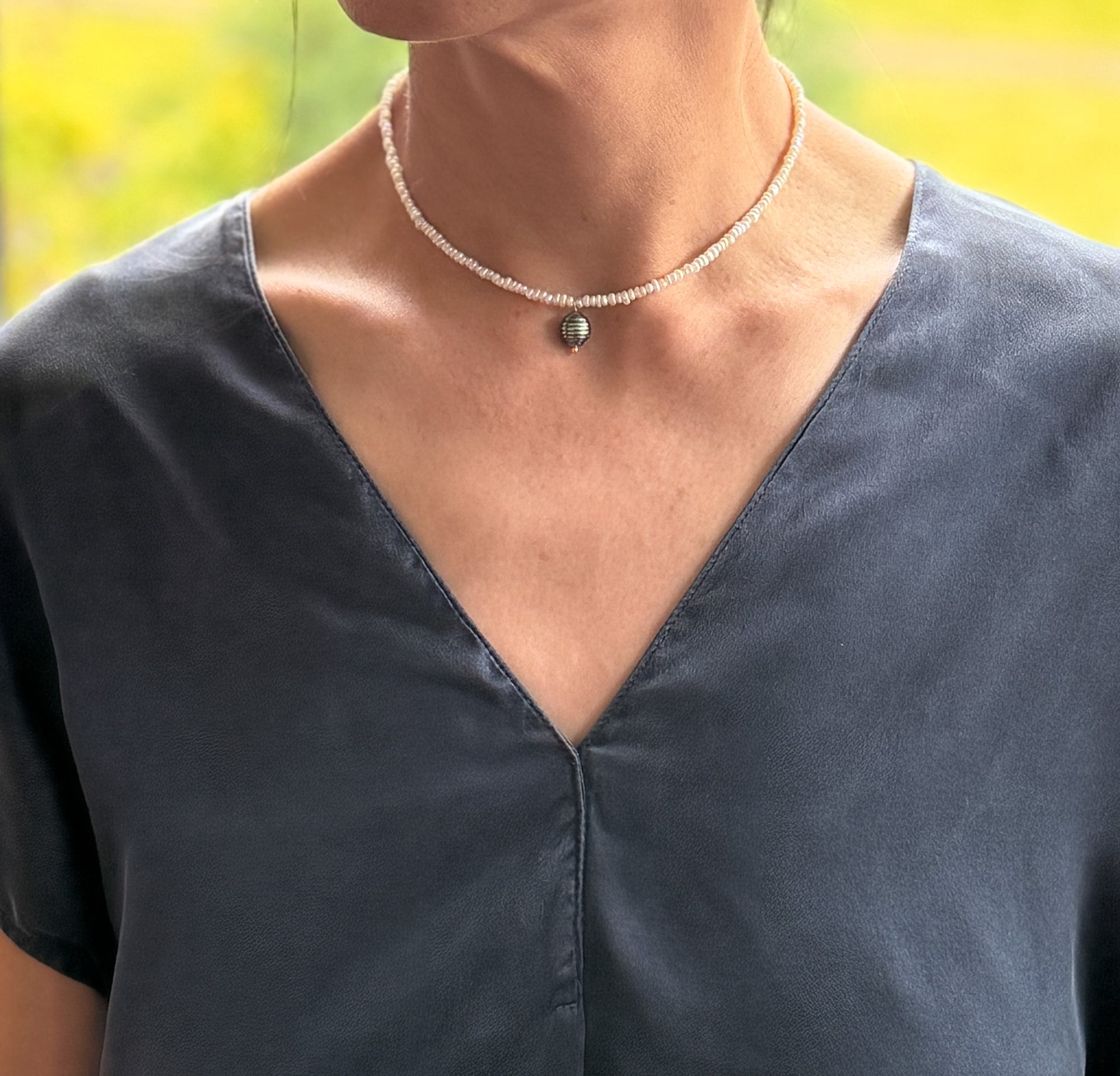 Model wearing white seed pearl choker with green Tahitian baroque pearl pendant over navy-blue v-neck top, showing the elegant drape and positioning at the collarbone with warm natural lighting