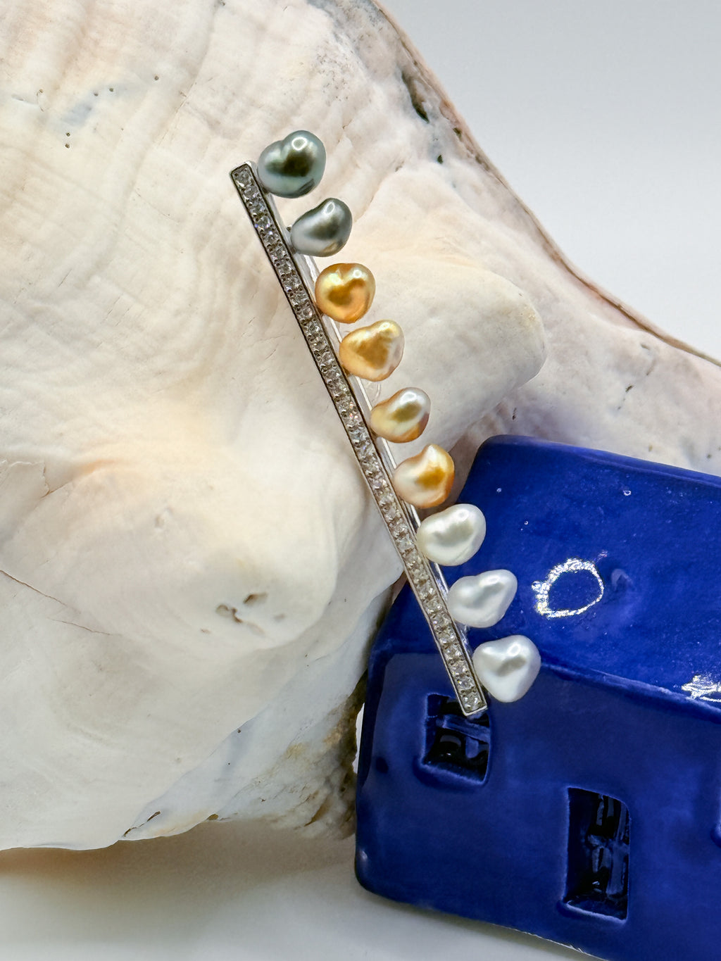 Close-up view of sterling silver bar pin brooch with heart-shaped keshi pearls on white conch shell and blue ceramic house, showcasing the beautiful variety of Tahitian and South Sea pearl colours in heart shapes