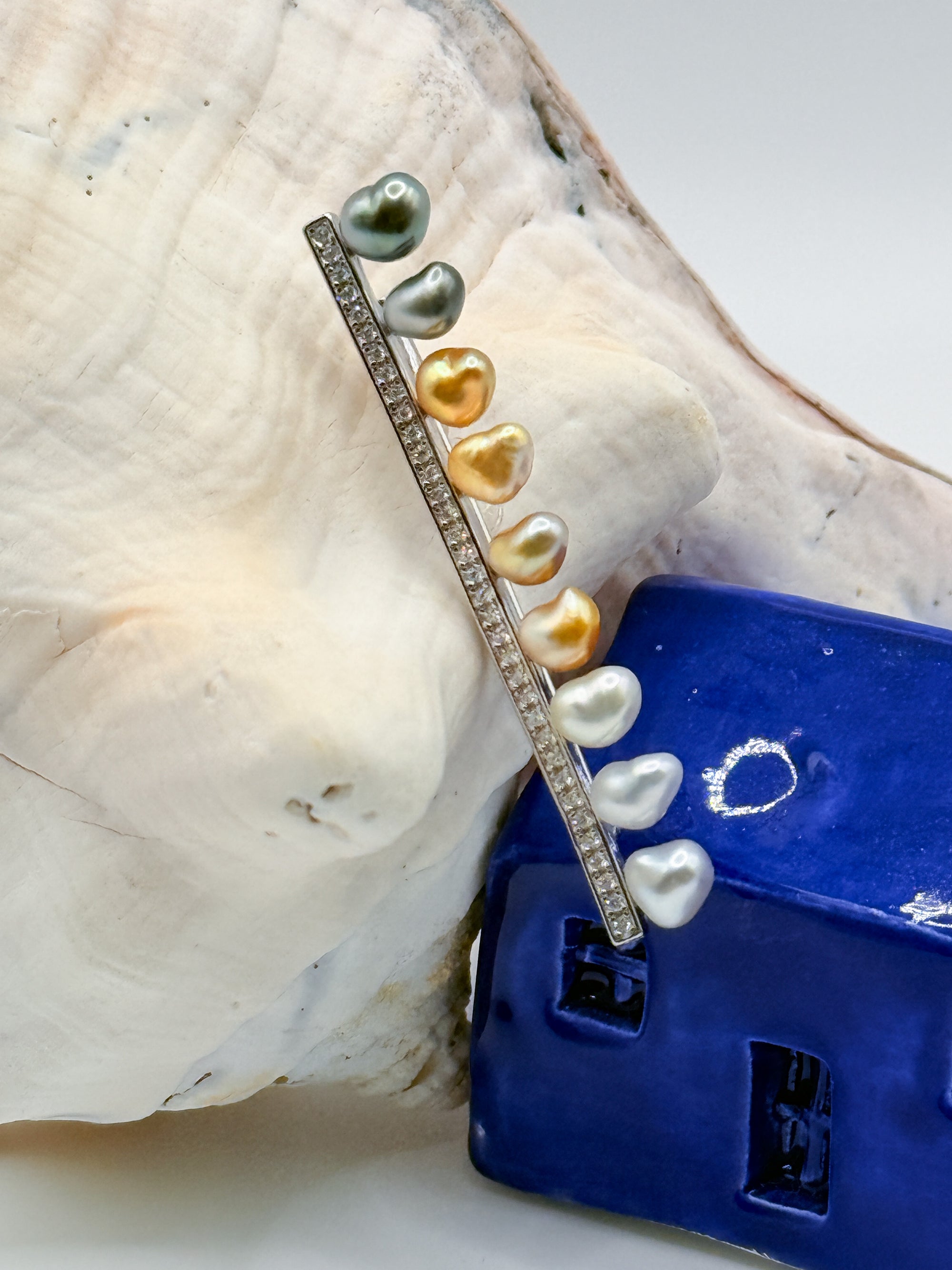 Close-up view of sterling silver bar pin brooch with heart-shaped keshi pearls on white conch shell and blue ceramic house, showcasing the beautiful variety of Tahitian and South Sea pearl colours in heart shapes