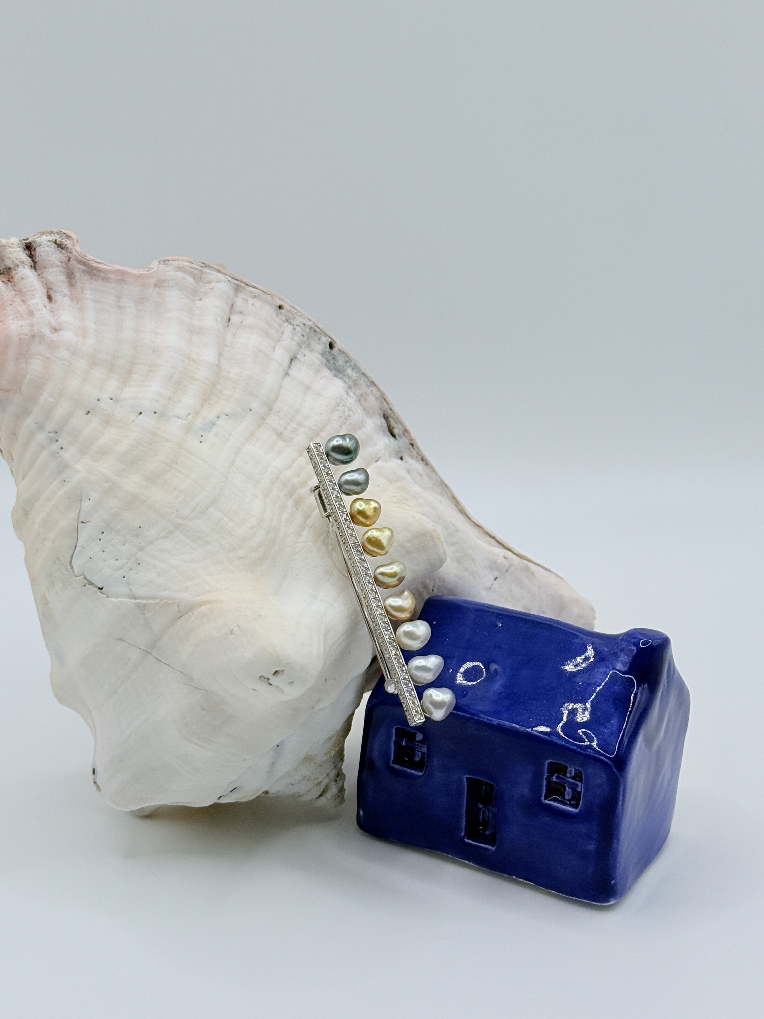 Artistic angle of sterling silver bar pin brooch with heart-shaped keshi pearls on white conch shell and blue ceramic house, emphasizing the crystal-studded bar and the beautiful gradient of pearl colours from dark green to golden to silver