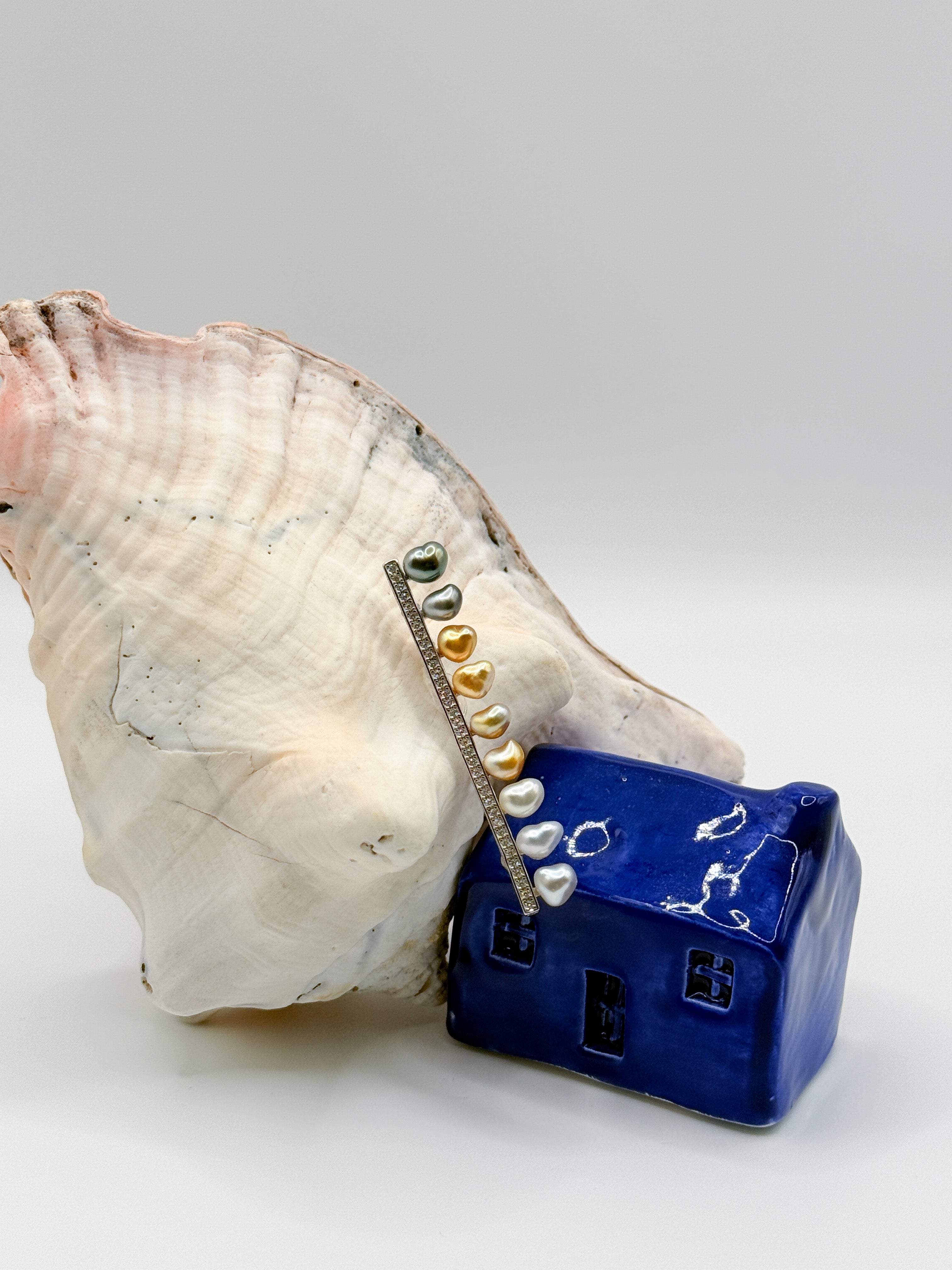 Sterling silver bar pin brooch with heart-shaped keshi pearls arranged on white conch shell next to blue ceramic house, showcasing the romantic heart shapes and diverse pearl tones