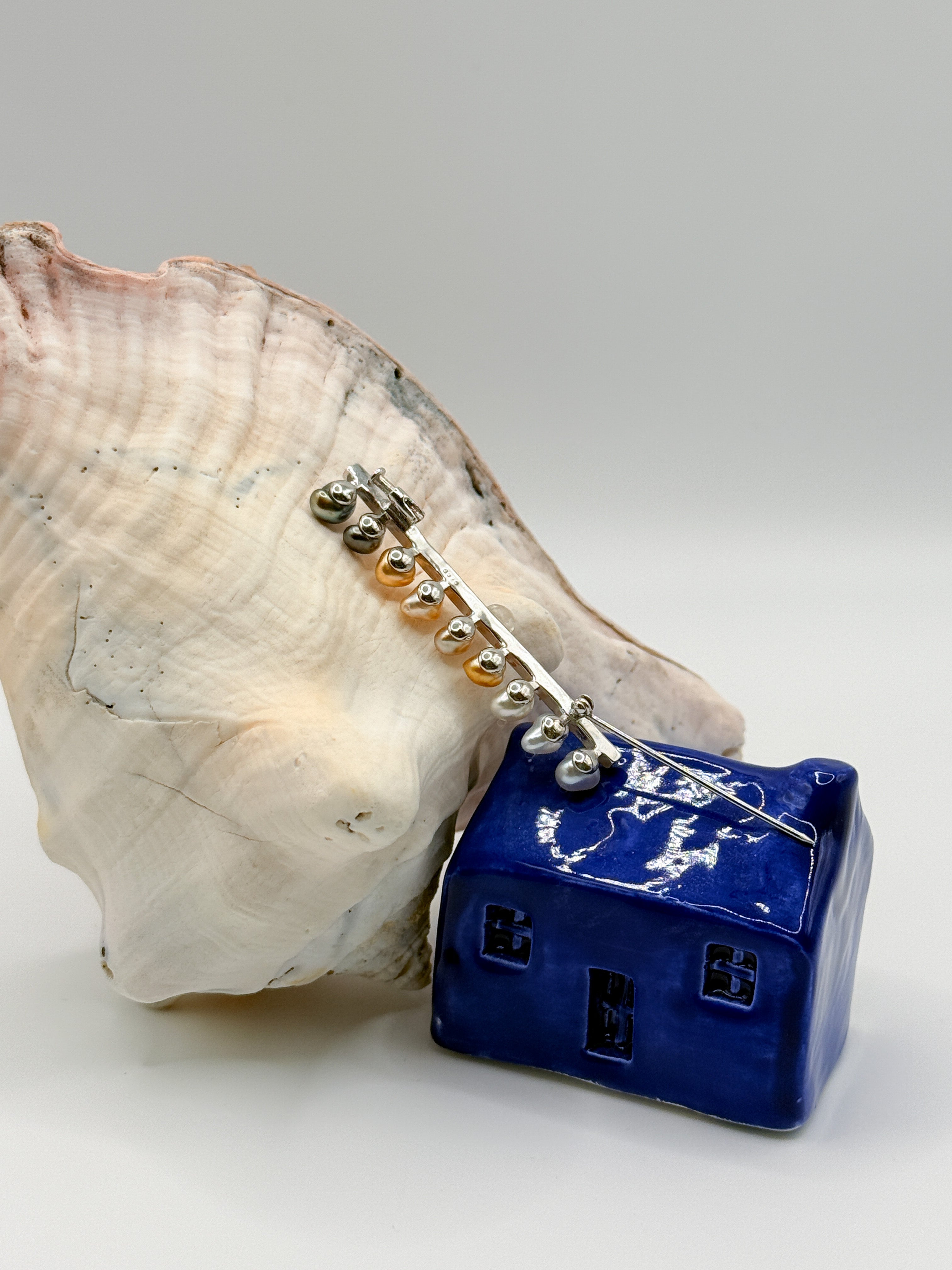 Sterling silver bar pin brooch with heart-shaped keshi pearls positioned on white conch shell and blue ceramic house, showing the detailed pin mechanism and gradient of pearl colours from green to golden to silver