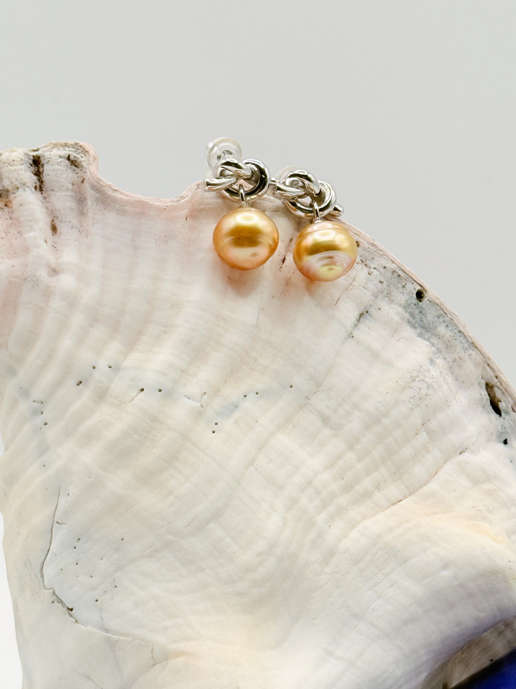 Close-up of sterling silver earrings with golden South Sea pearls and decorative knot details positioned on white conch shell, showcasing the lustrous golden pearl surfaces and detailed silver craftsmanship
