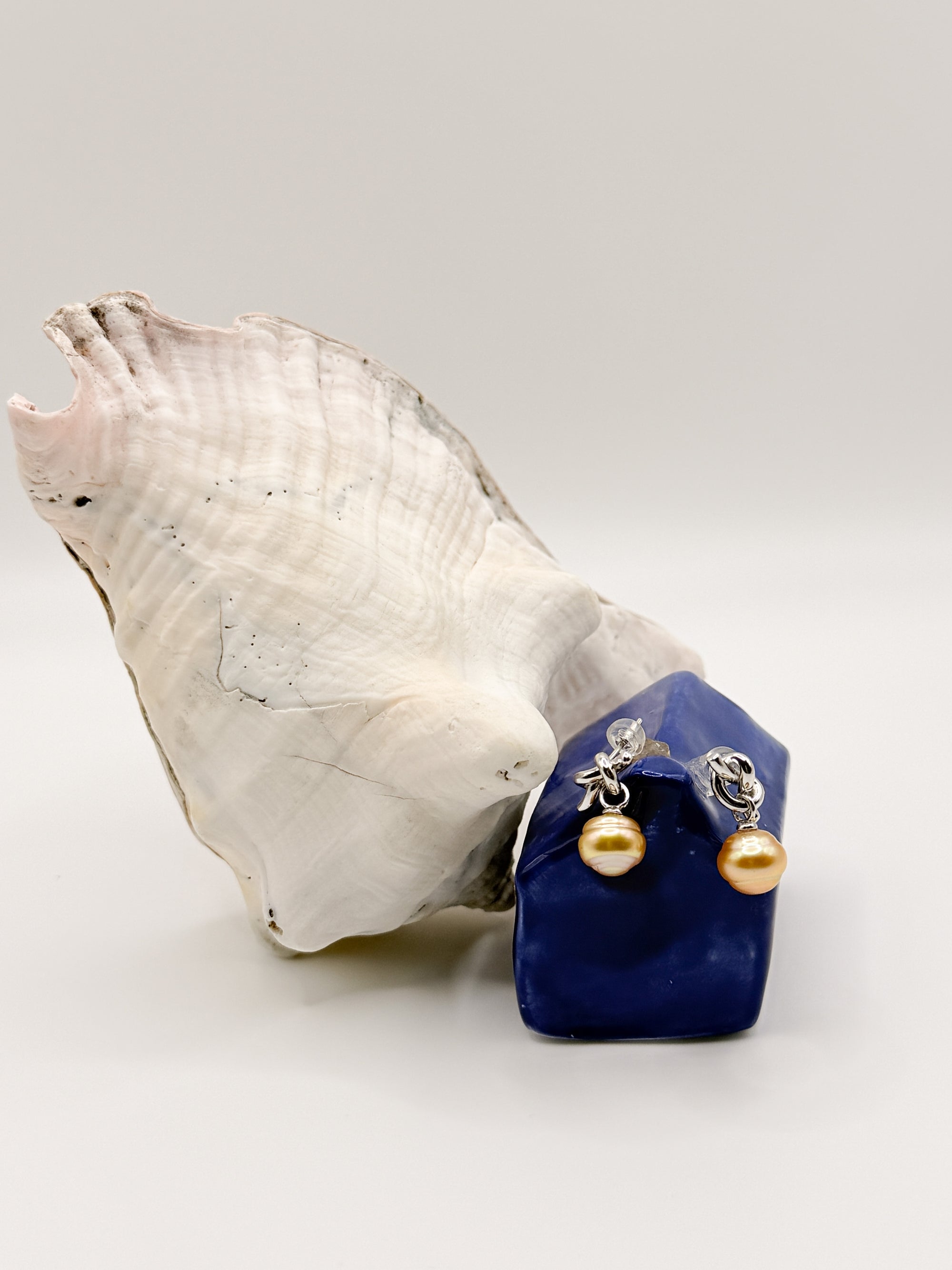 Sterling silver earrings with golden South Sea pearls positioned on blue ceramic house next to white conch shell, highlighting the warm golden pearl tones and intricate silver knot work