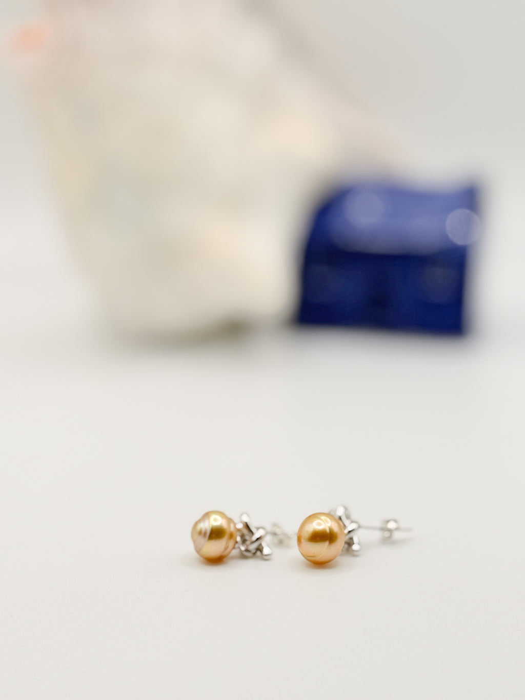 Golden South Sea pearl earrings with sterling silver knot details laid flat on white background with soft focus shell and blue ceramic house in background