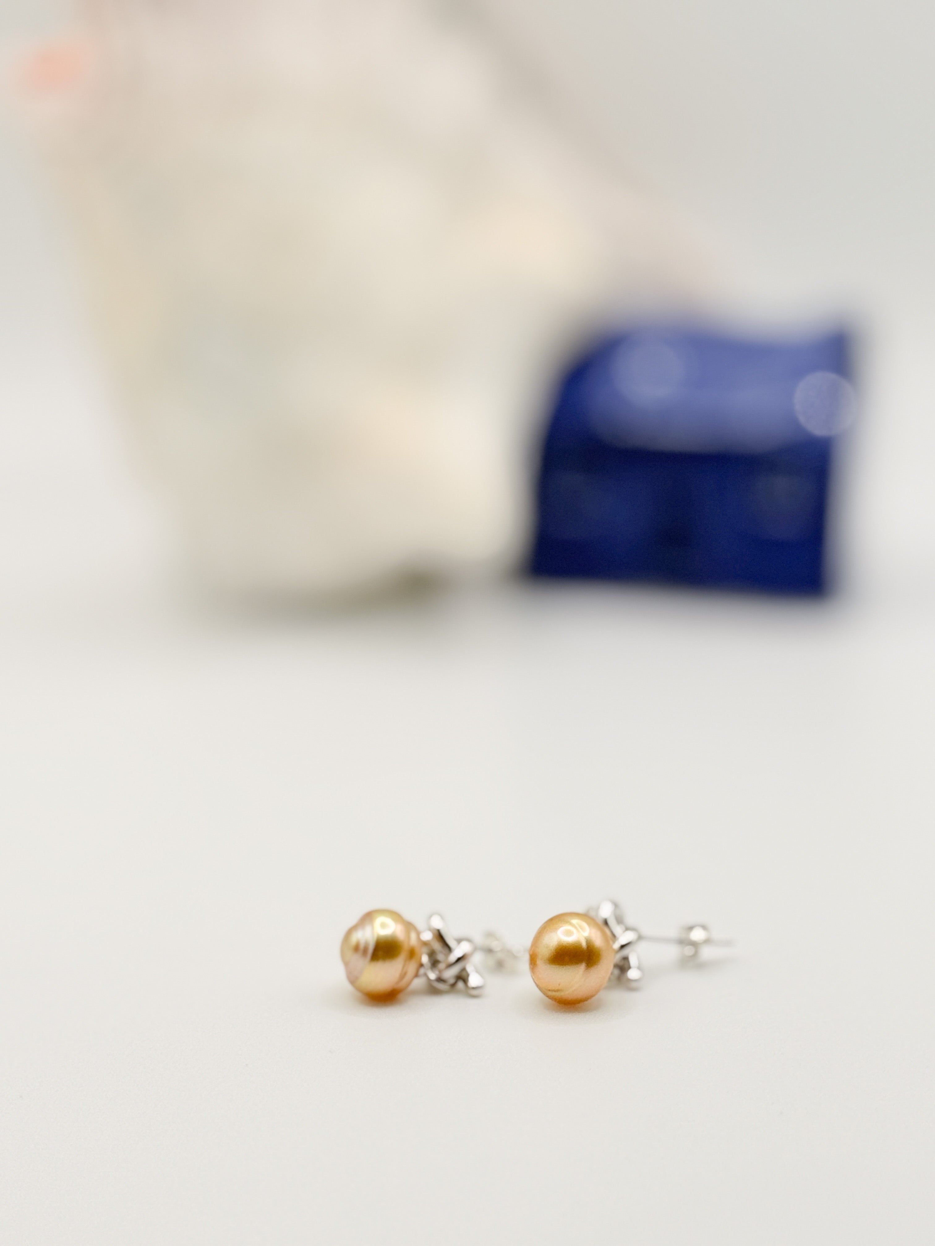 Golden South Sea pearl earrings with sterling silver knot details laid flat on white background with soft focus shell and blue ceramic house in background