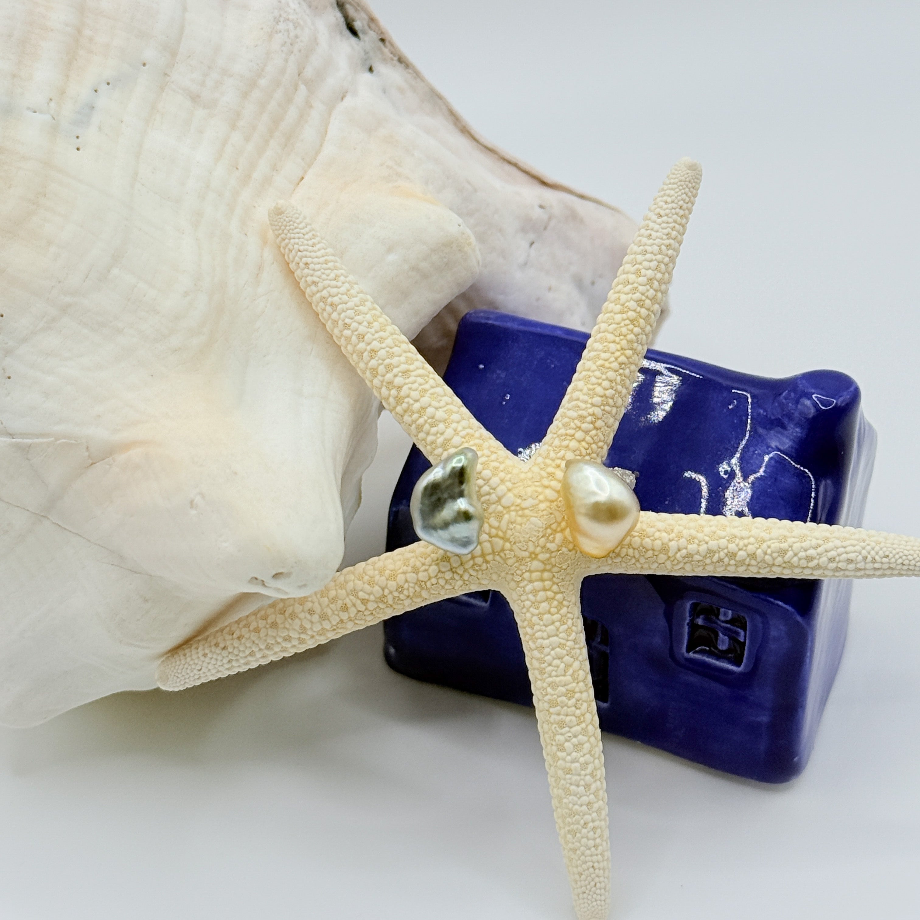 Half-moon Tahitian gold South Sea pearl stud earrings asymmetrical baroque pearls crescent moon shaped ear studs resting on starfish against blue house and seashell