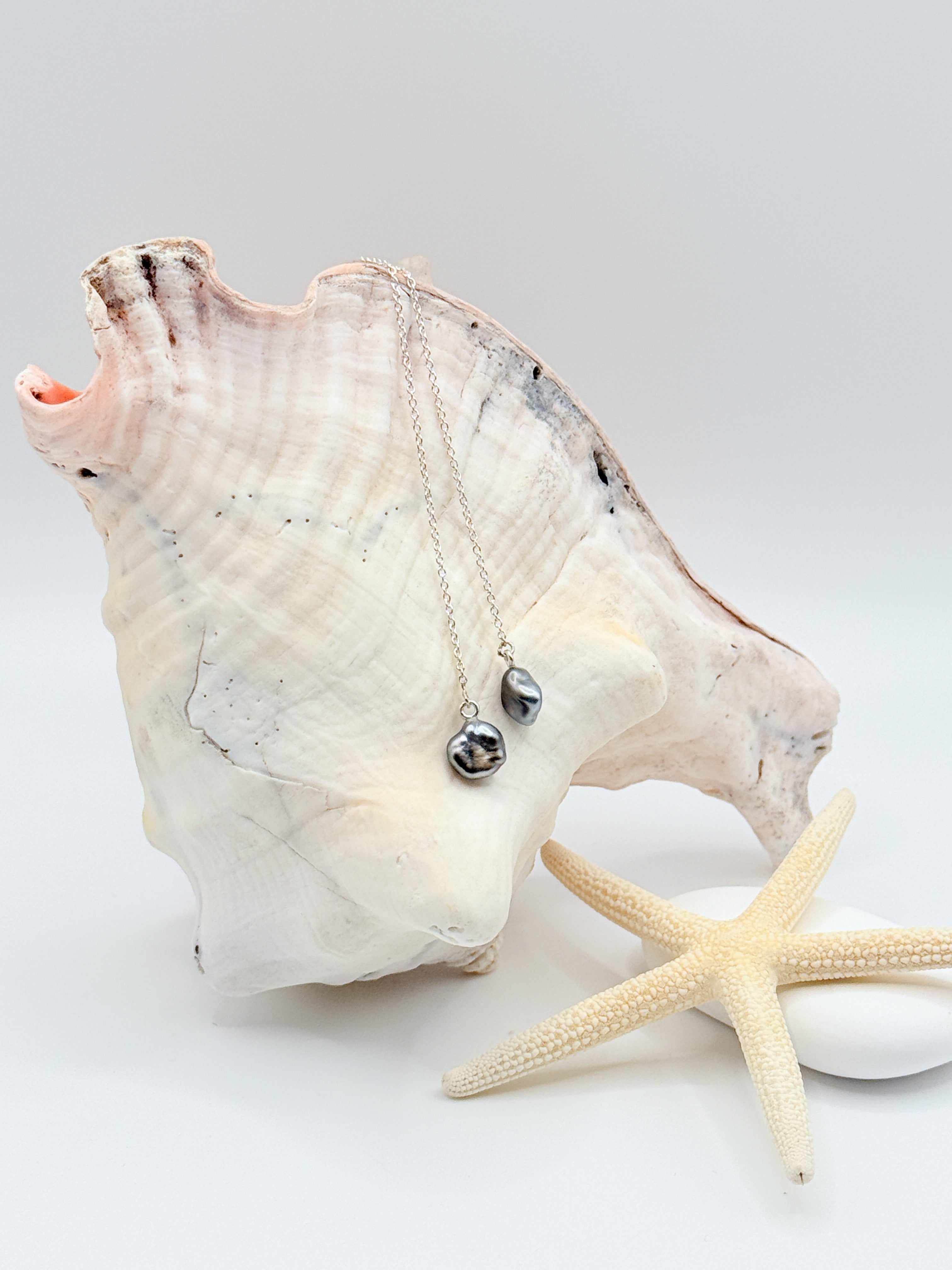 Tahitian Keshi pearl threader earrings showing dark silver pearls green slate hints sterling silver chains draped over conch and starfish