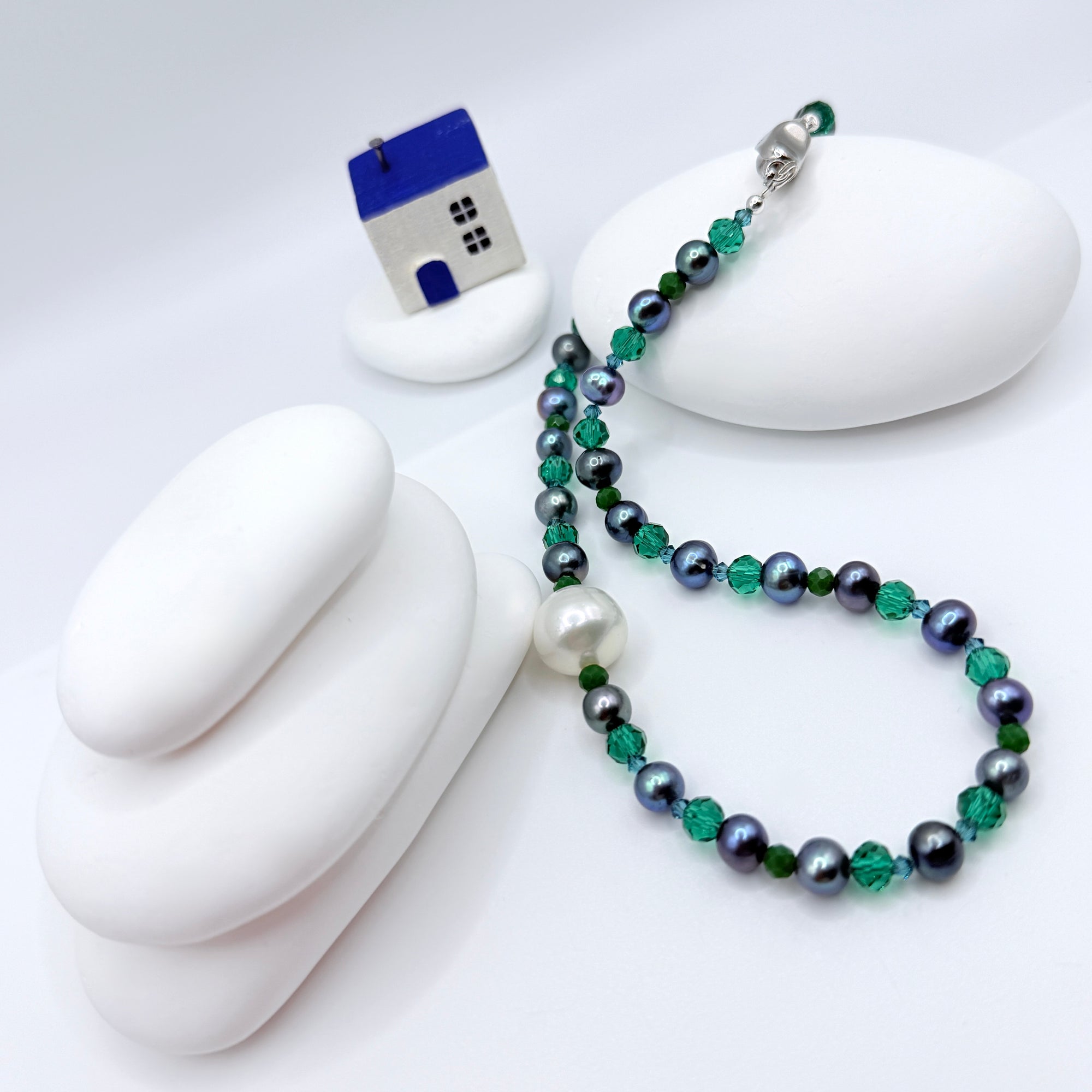 Necklace with white south sea baroque pearl with blude dye-coloured freshwater pearls and green glass beads on a white surface with a small house model.