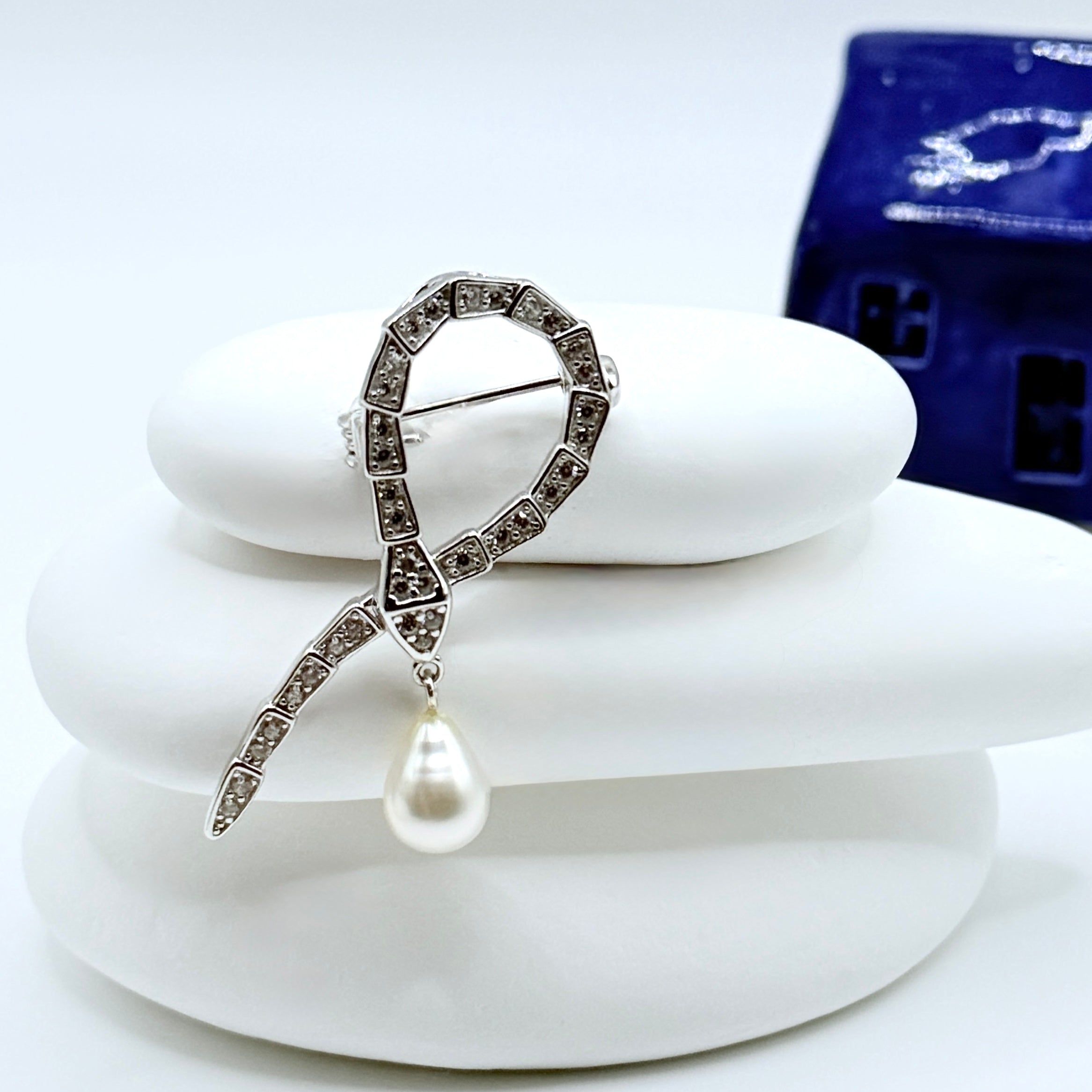 Snake shape sterling silver brooch pendant with white South Sea Keshi pearl drop and CZ stones displayed on white stones with blue ceramic house