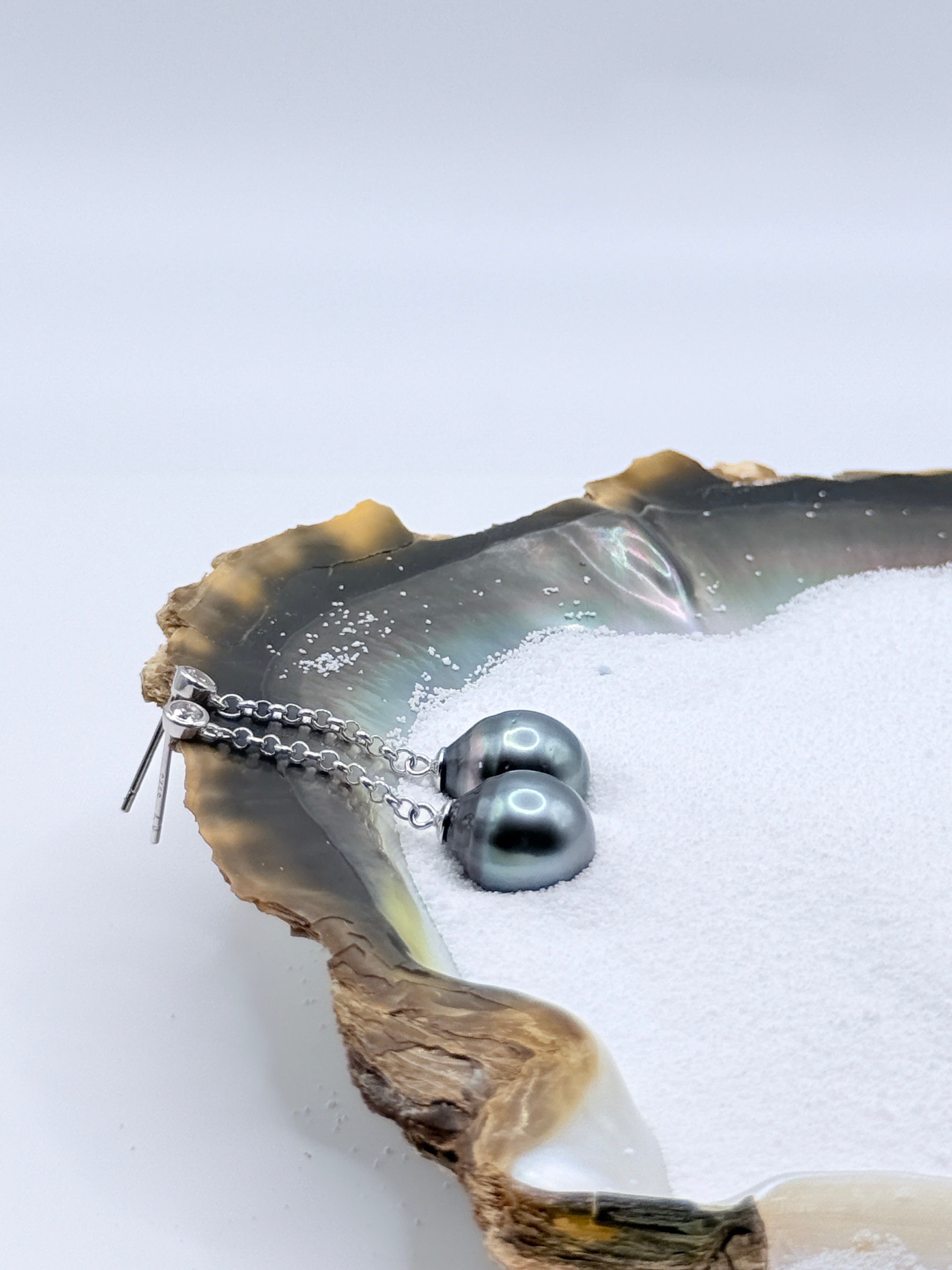 Close-up view of sterling silver chain earrings with CZ bezels and charcoal grey Tahitian baroque pearls in oyster shell highlighting silvery lustre