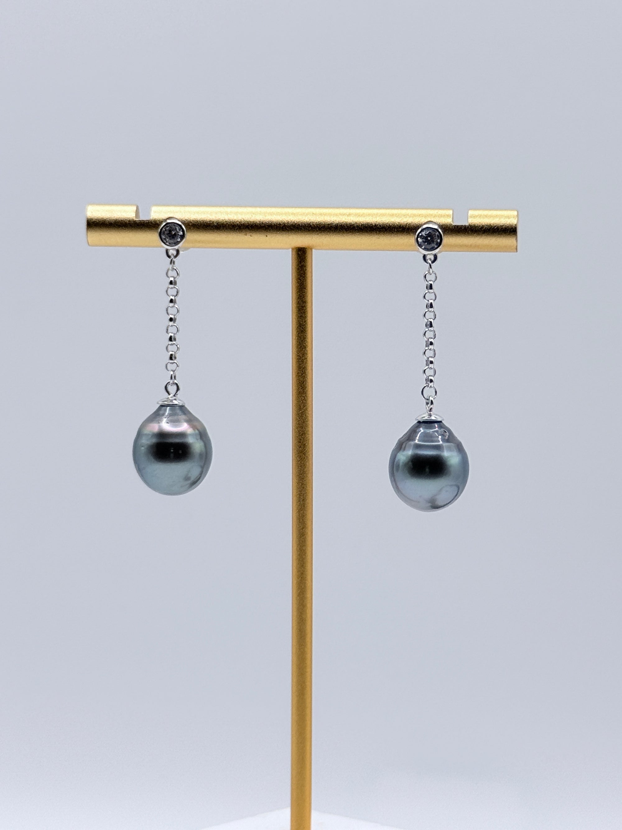 Tahitian baroque pearl drop earrings with sterling silver CZ bezel chains displayed on gold earring stand showing dark grey pearls with metallic overtones