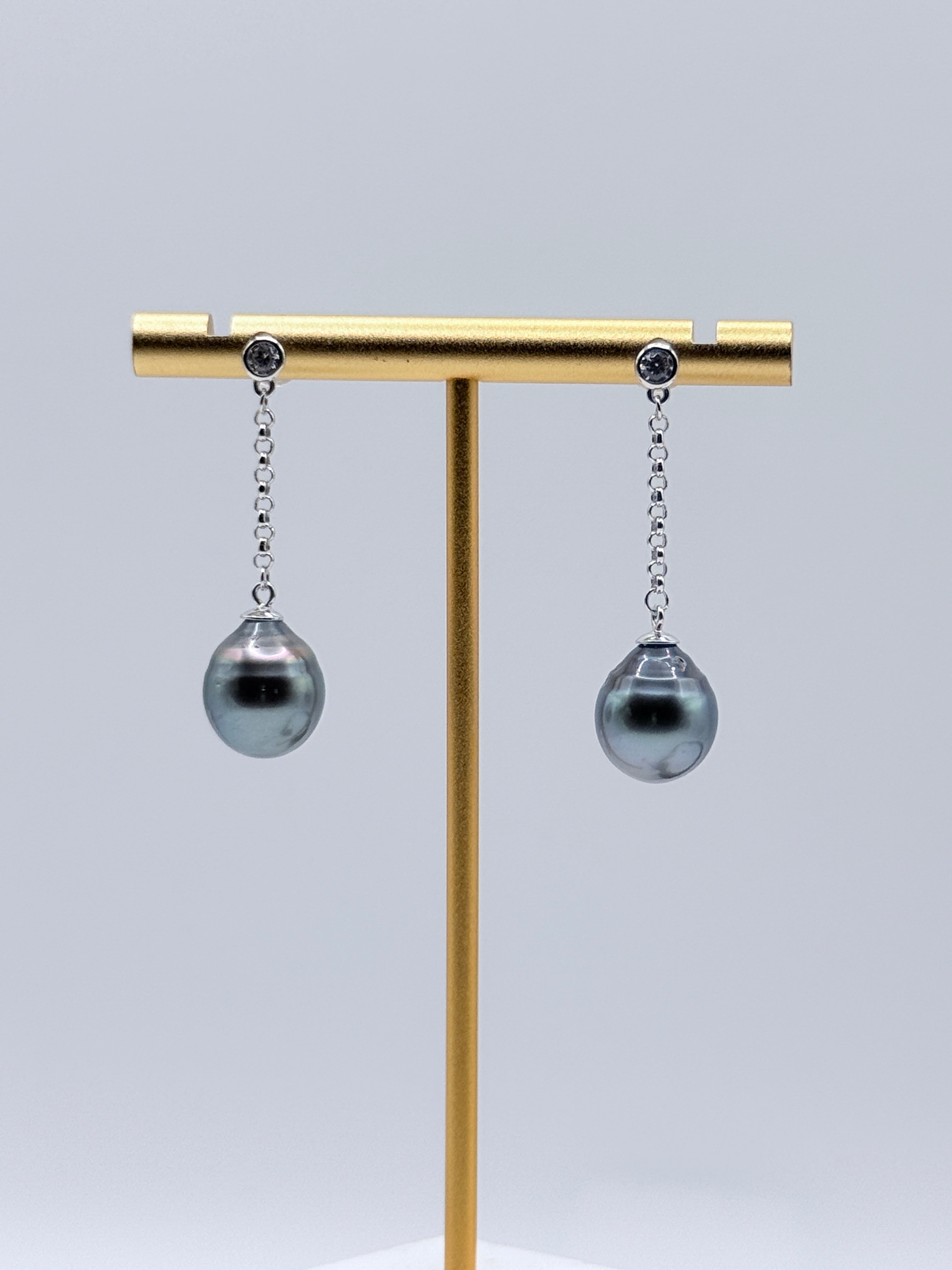 Tahitian baroque pearl drop earrings with sterling silver CZ bezel chains displayed on gold earring stand showing dark grey pearls with metallic overtones