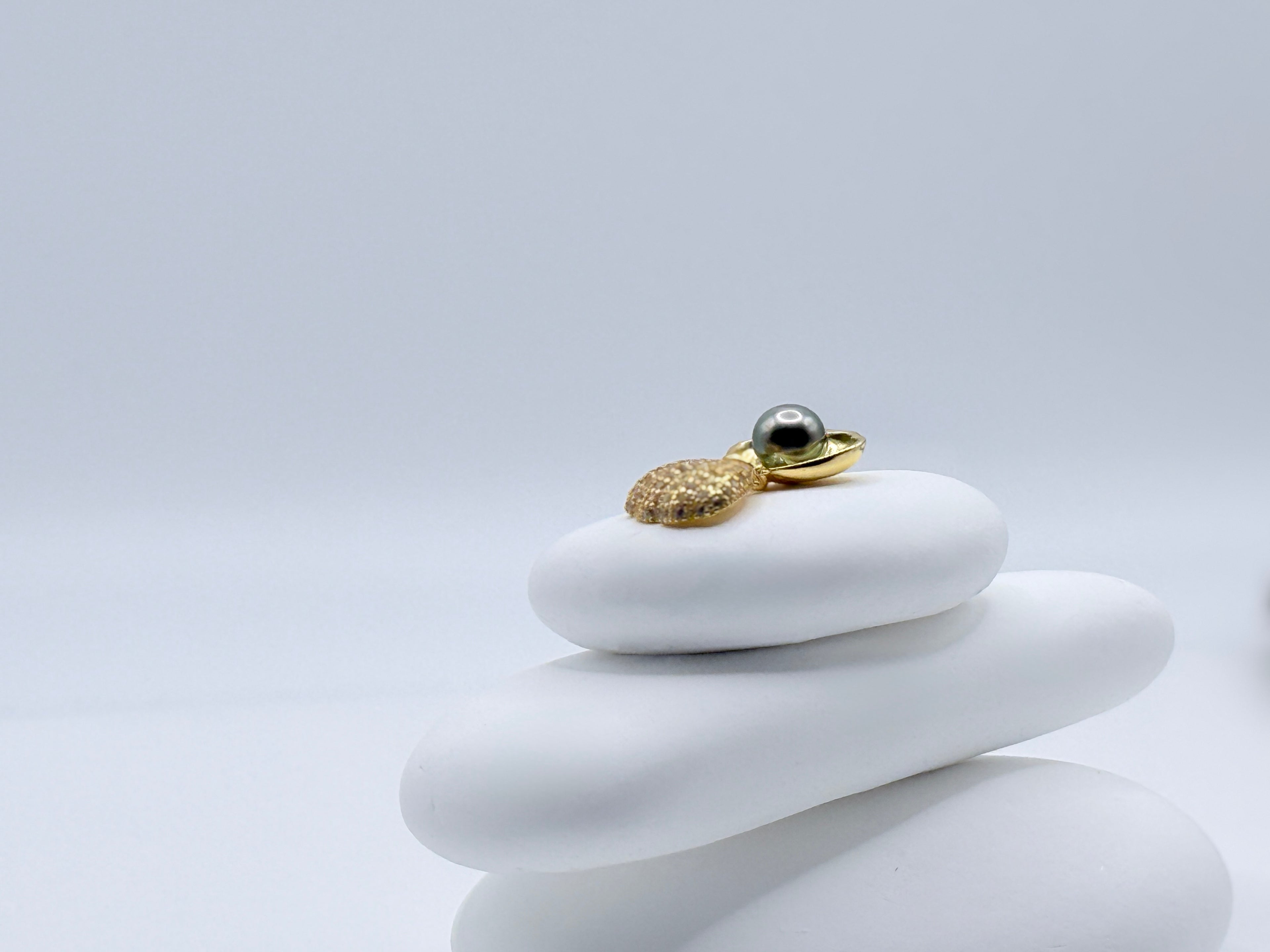 Sea Shell Serenade gold plated scallop shell pendant with dark grey 7mm Tahitian keshi pearl displayed on white stone stack showing ocean-inspired design