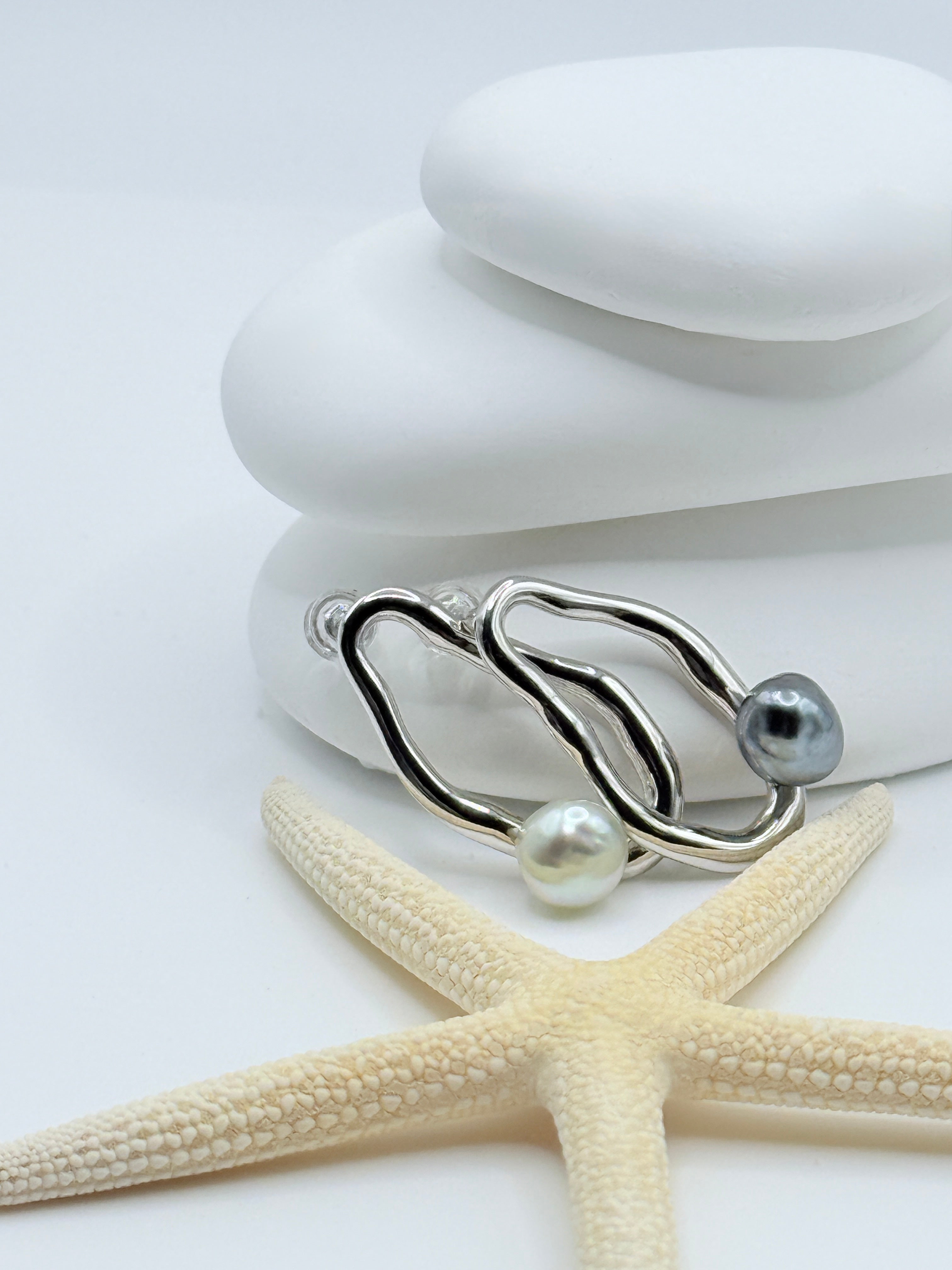 Pebble asymmetric earrings displaying flowing sterling silver forms with contrasting keshi pearls on white stone stack with starfish styling
