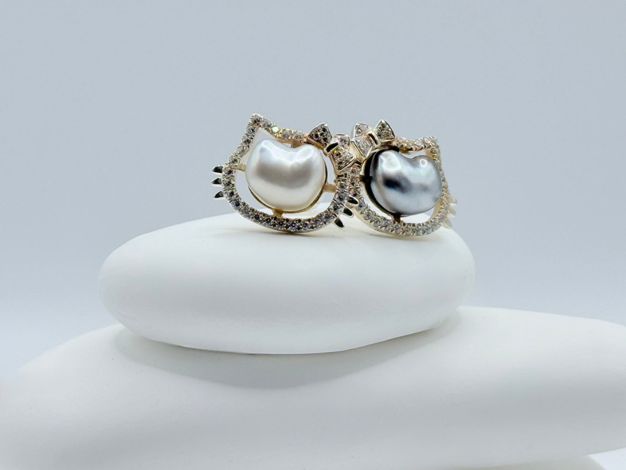 Close-up view of Hello Kitty sterling silver ear studs with white and dark grey keshi pearls surrounded by sparkling CZ halos