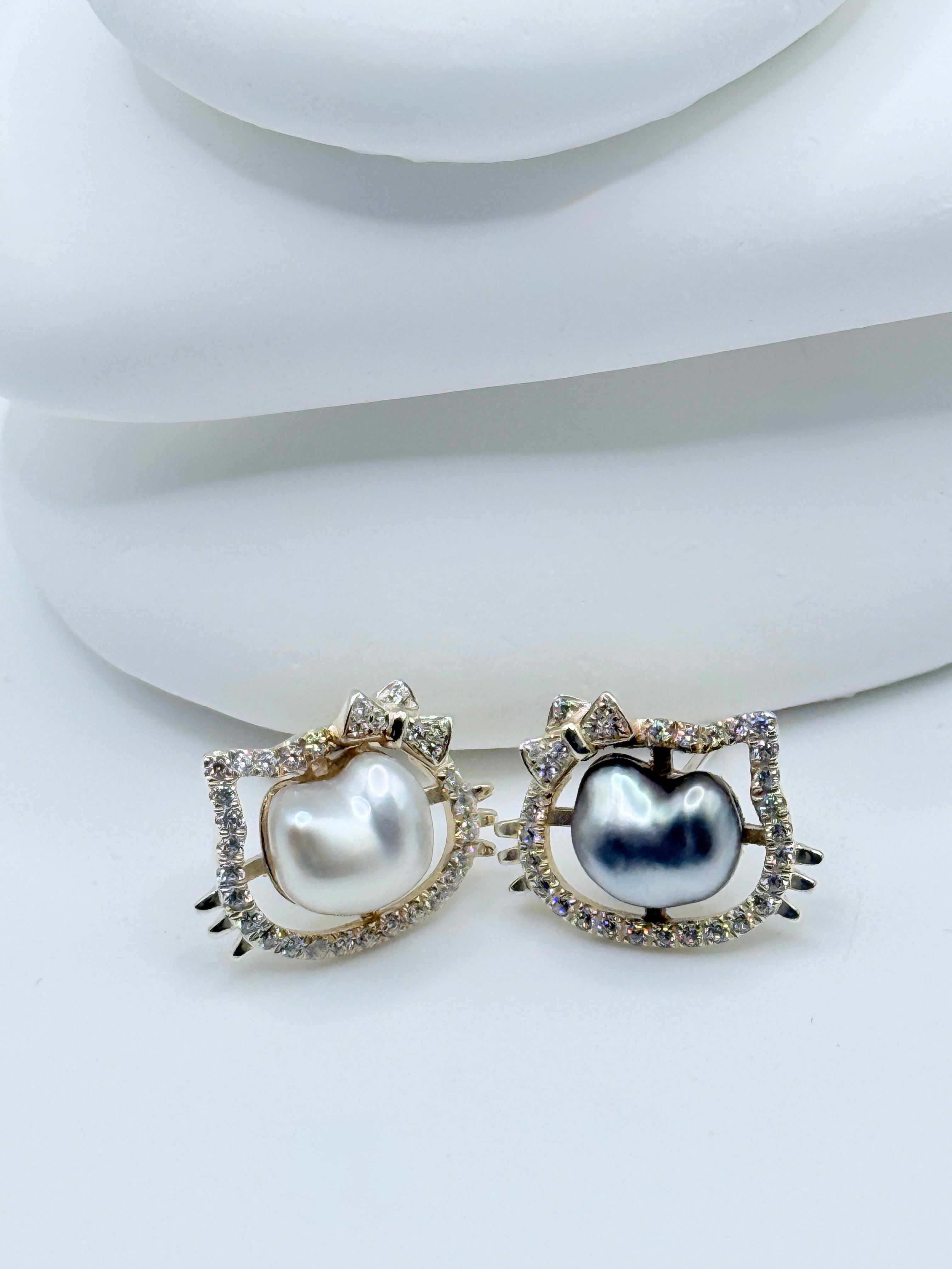 Sterling silver Hello Kitty ear studs with CZ halos and contrasting keshi pearls displayed on white curved surfaces