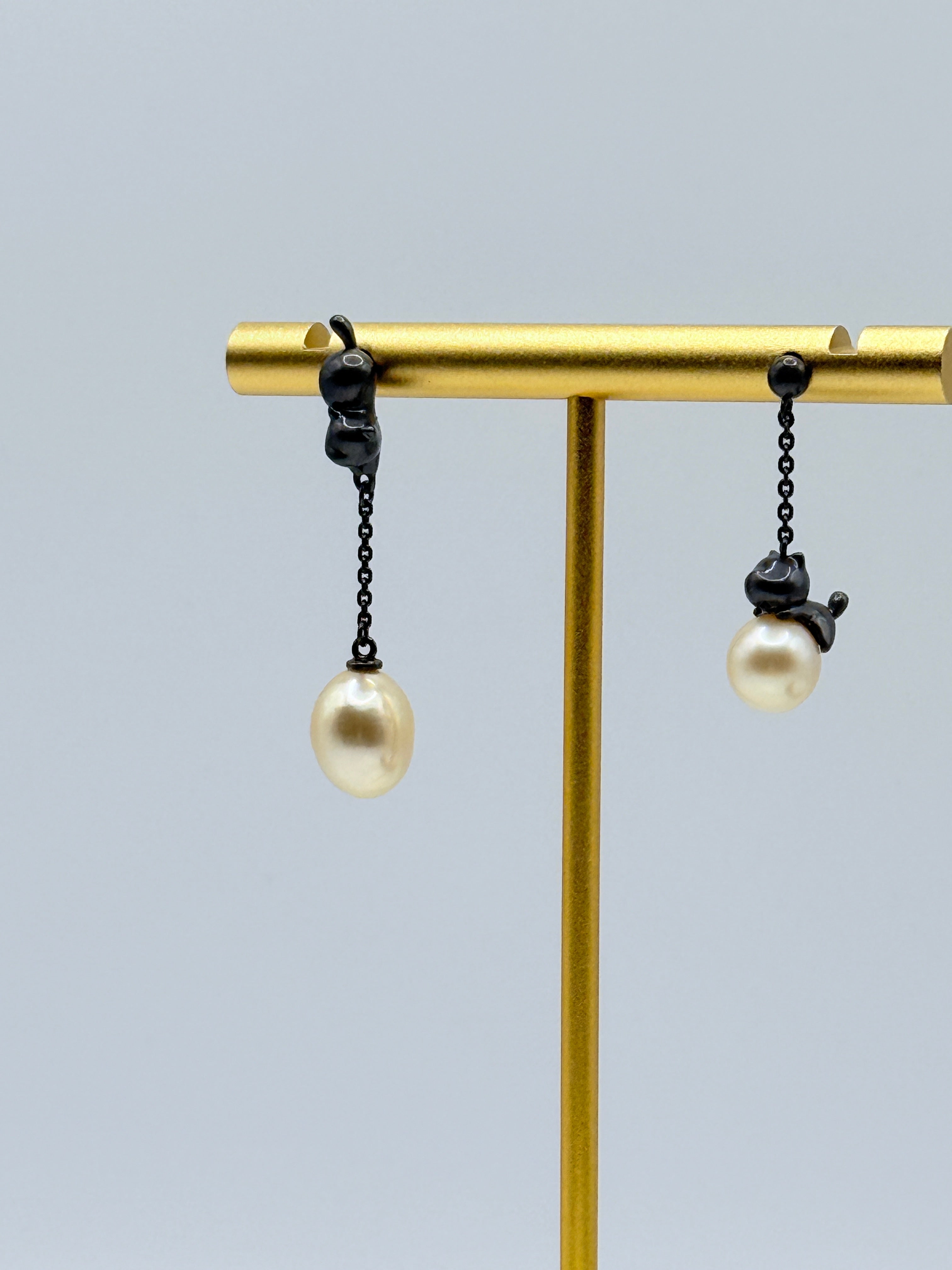 Complete set of black rhodium plated sterling silver cat dangle earrings with light gold keshi pearls displayed on gold earring stand