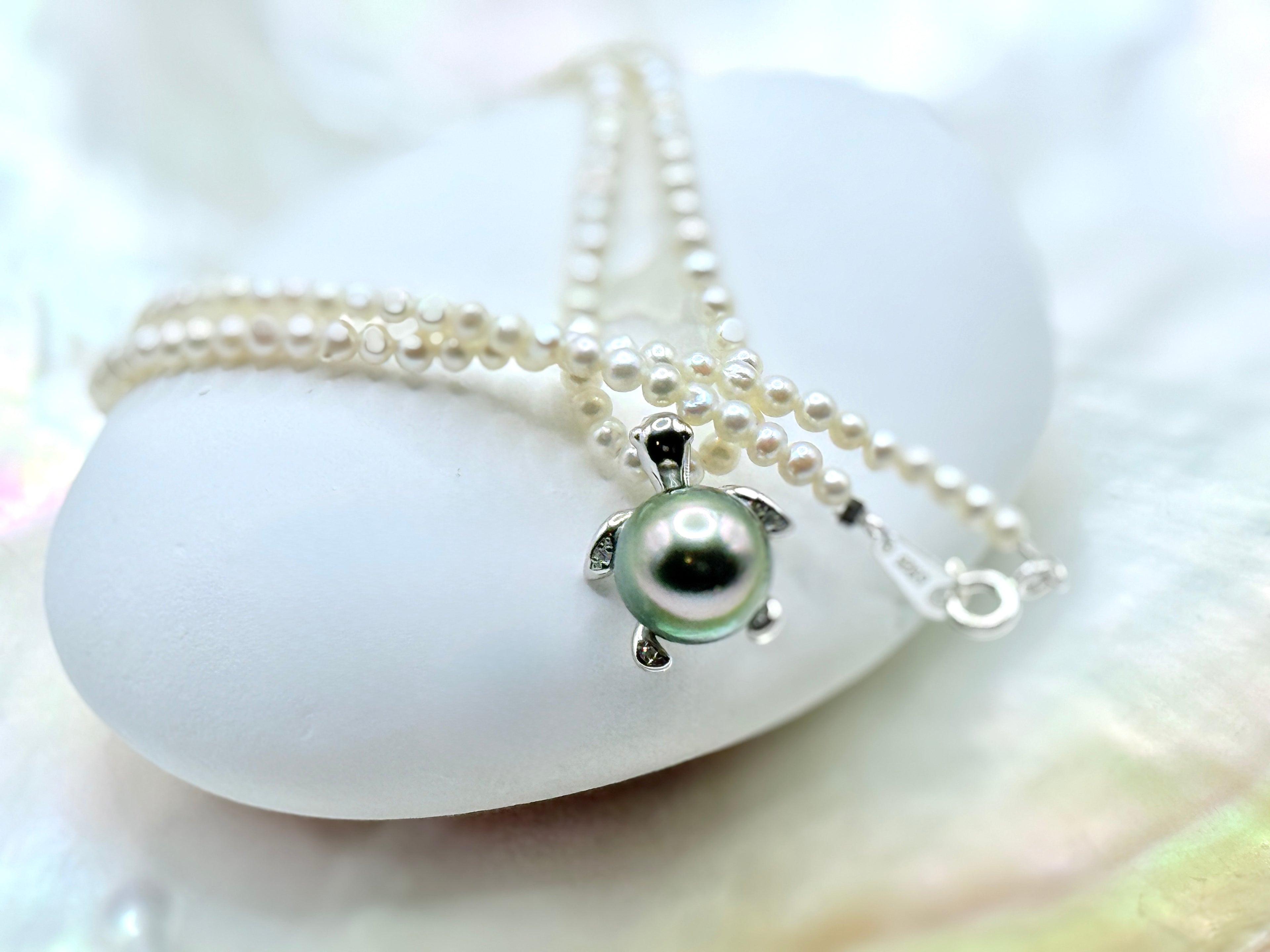 Close-up of 10mm green Tahitian pearl in sea turtle pendant setting with 2.5mm white seed pearl strand on white shell