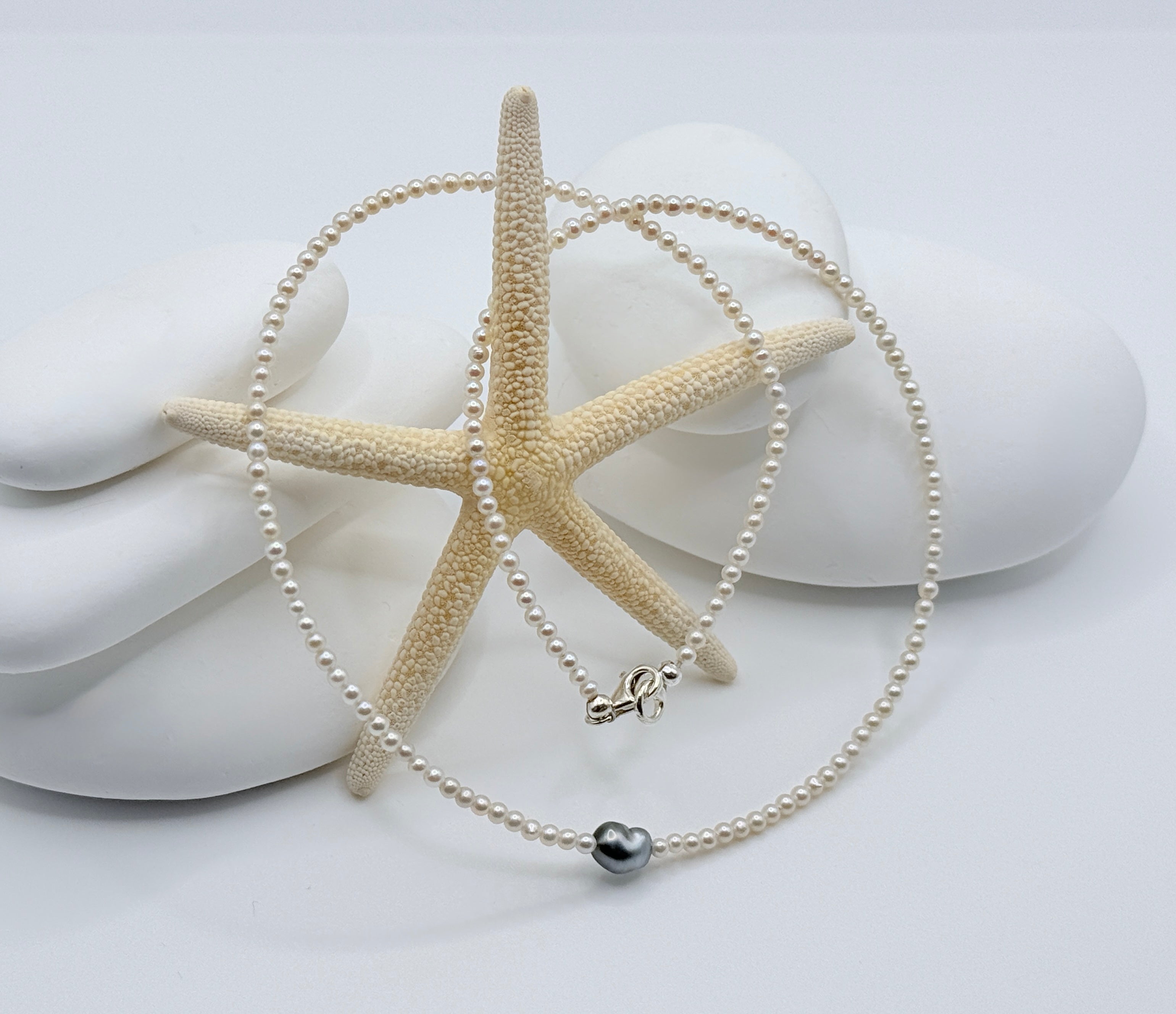 White freshwater seed pearl strand with heart-shaped Tahitian keshi pendant and sterling silver clasp on white shells and starfish