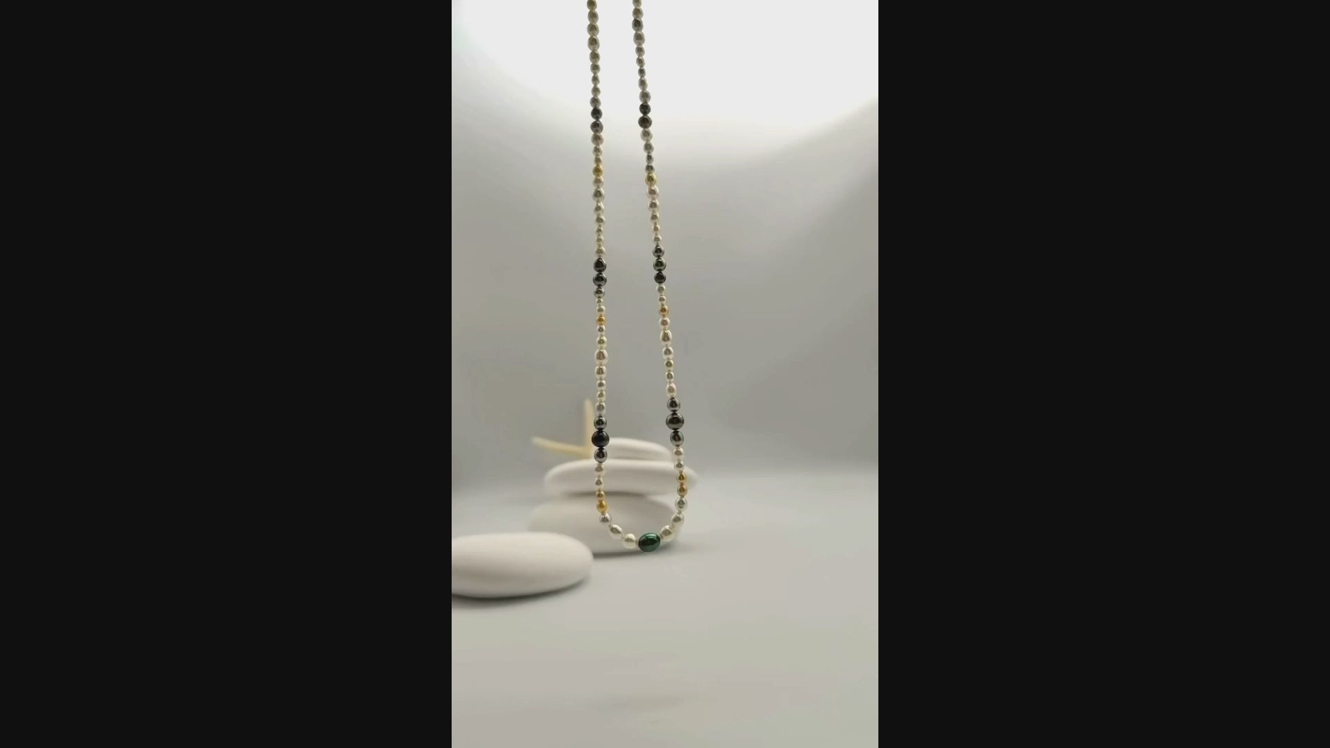 Multi-coloured pearl strand necklace suspended and gently moving in the air, displaying the beautiful variety of white, golden, silver, and dark green pearls in fluid motion