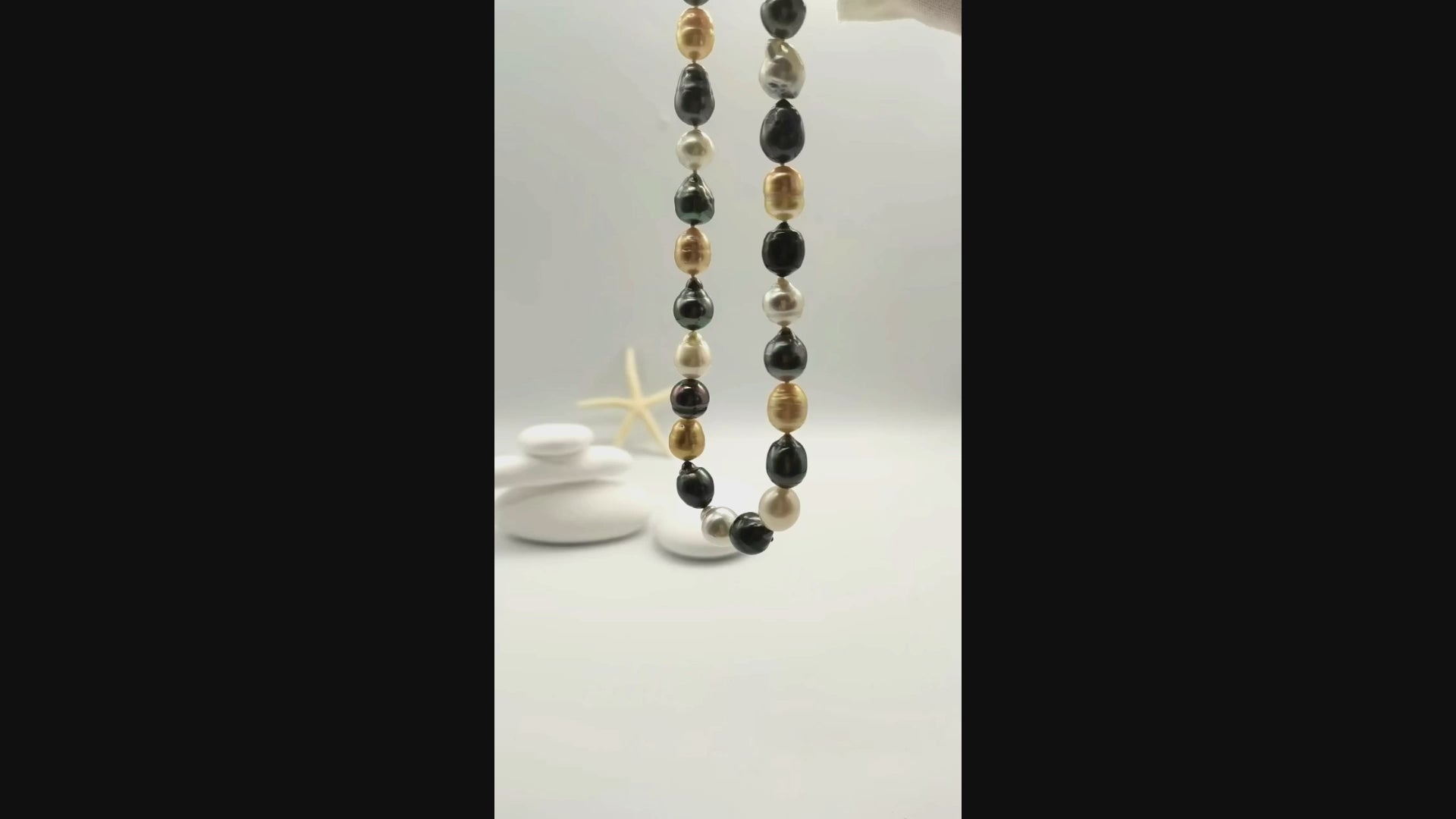 Multicoloured baroque pearl necklace suspended and gently moving in the air, displaying the beautiful variety of white, golden, dark green and black baroque pearls in fluid motion